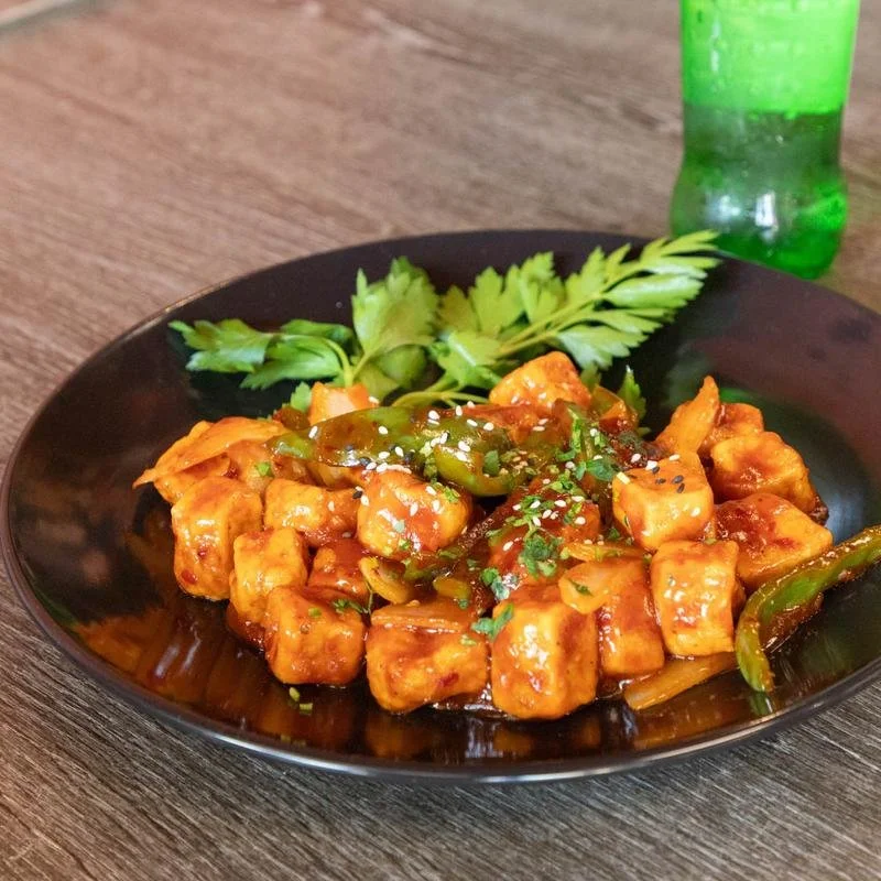 A black bowl of spicy orange chicken with green peppers and a sprig of cilantro on top, served on a wooden table, with a green bottle in the background.