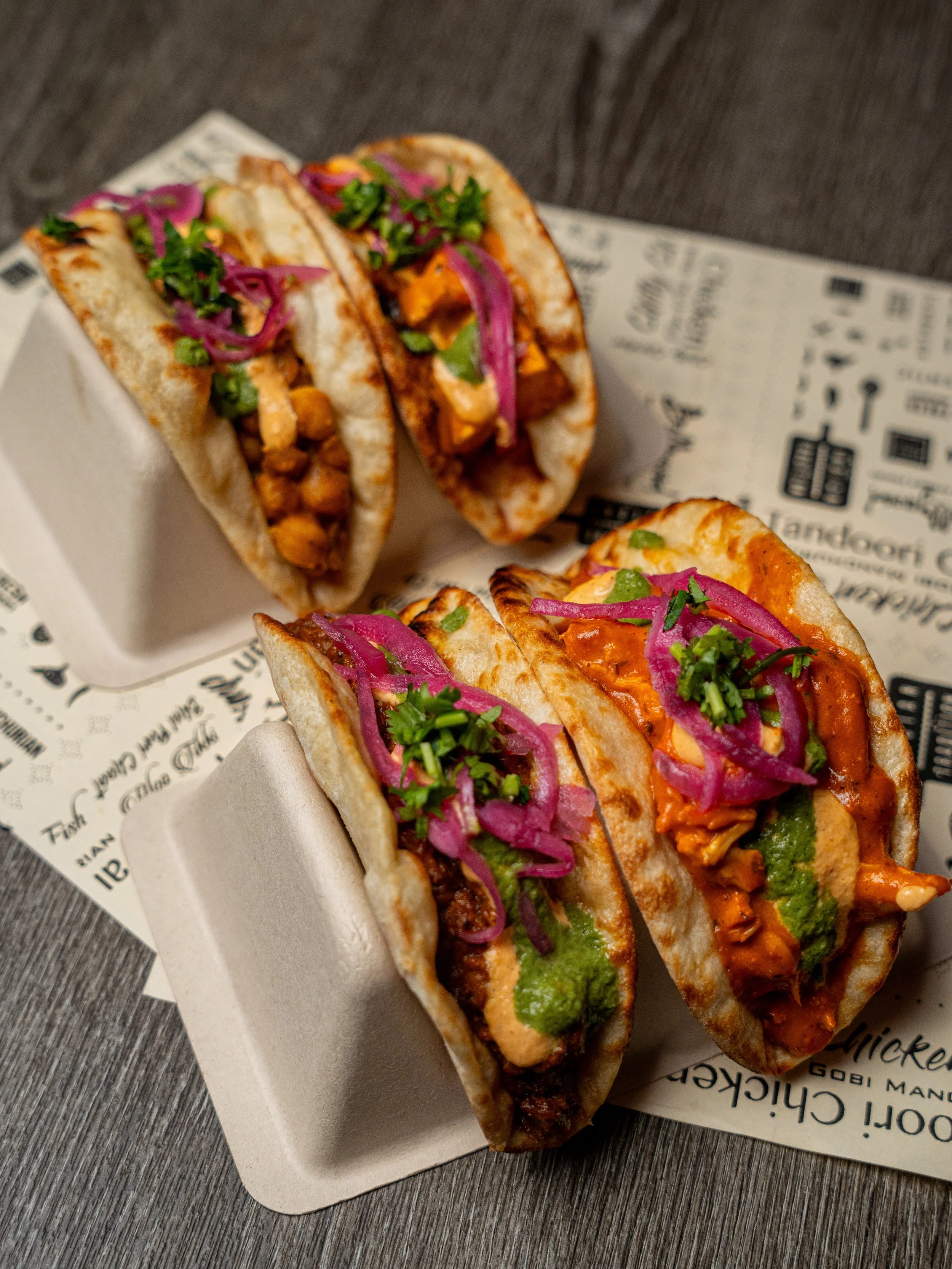 Three tacos filled with chicken, topped with chopped cilantro and pickled red onions, served on a white paper tray with printed branding, placed on a dark wooden surface.