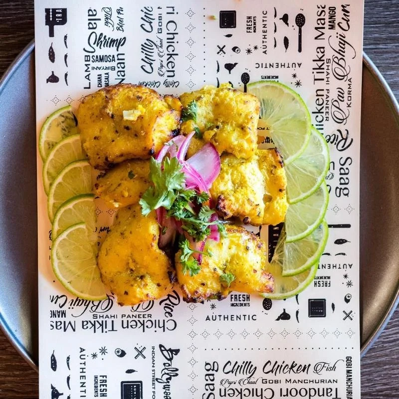 Four pieces of grilled or baked fish topped with yellow sauce on a white paper-lined plate. Sliced lime wedges surround the fish, garnished with a sprig of cilantro and sliced red onion. The paper has black text and icons related to food and cooking.