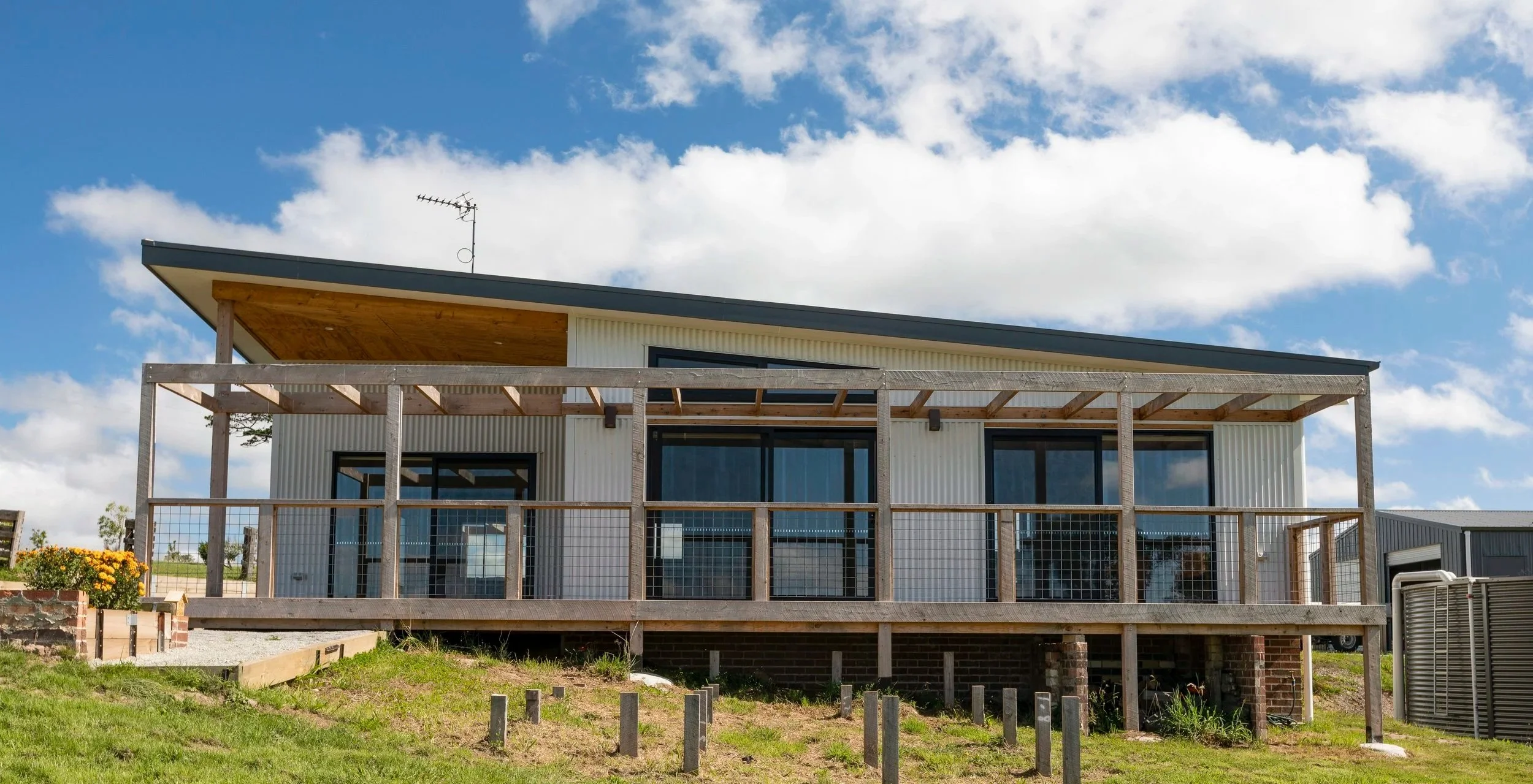 A modern house under construction with a wooden deck in front, large glass windows, a sloped roof, and an outdoor antenna on top, against a partly cloudy sky.