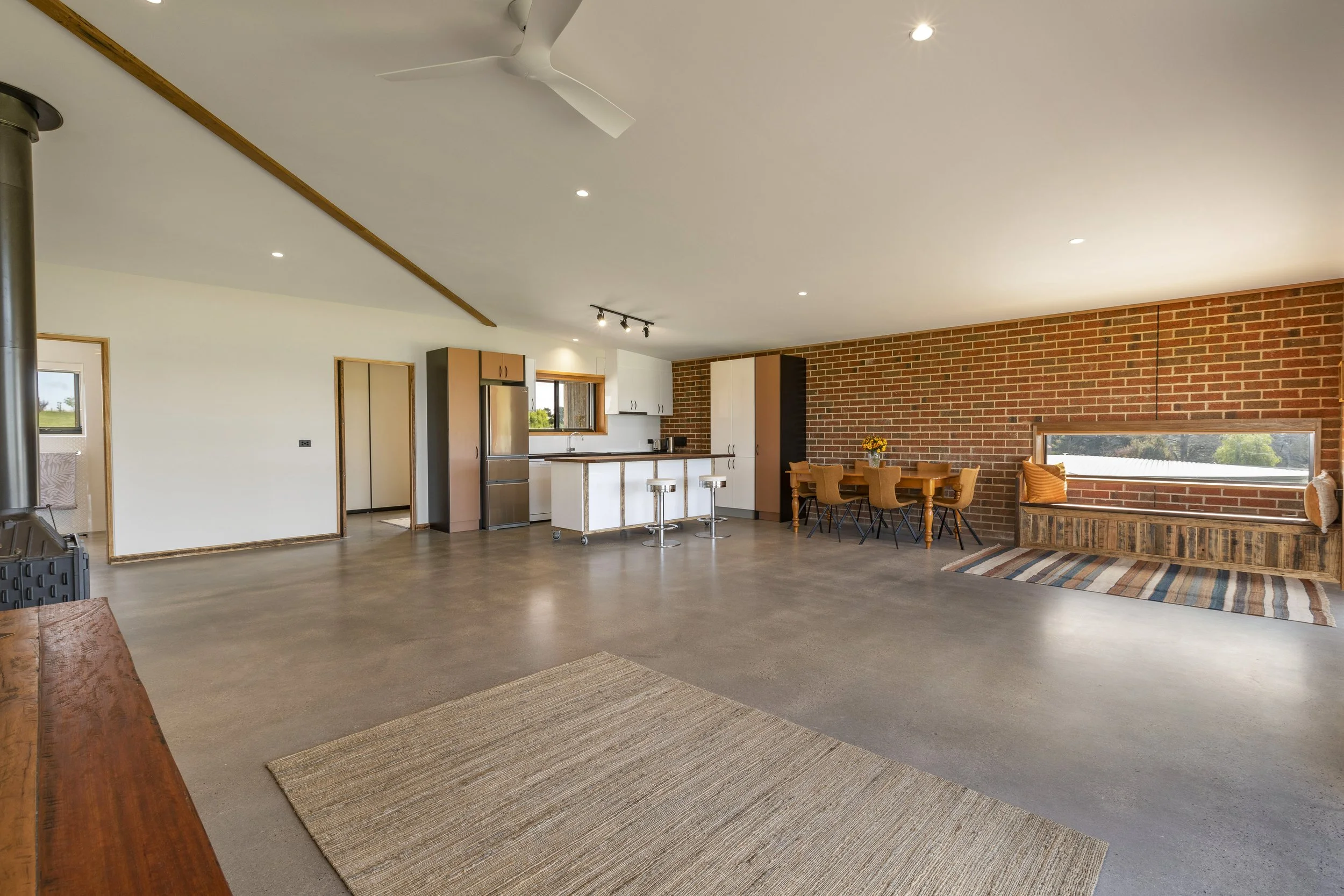 A modern open-concept kitchen and dining area with brick accent wall, large windows, wooden furniture, and minimal decor.