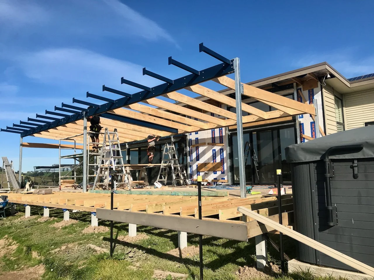 Construction of a patio or deck extension on a house with workers on ladders installing wooden beams and framework, with blue sky in the background.