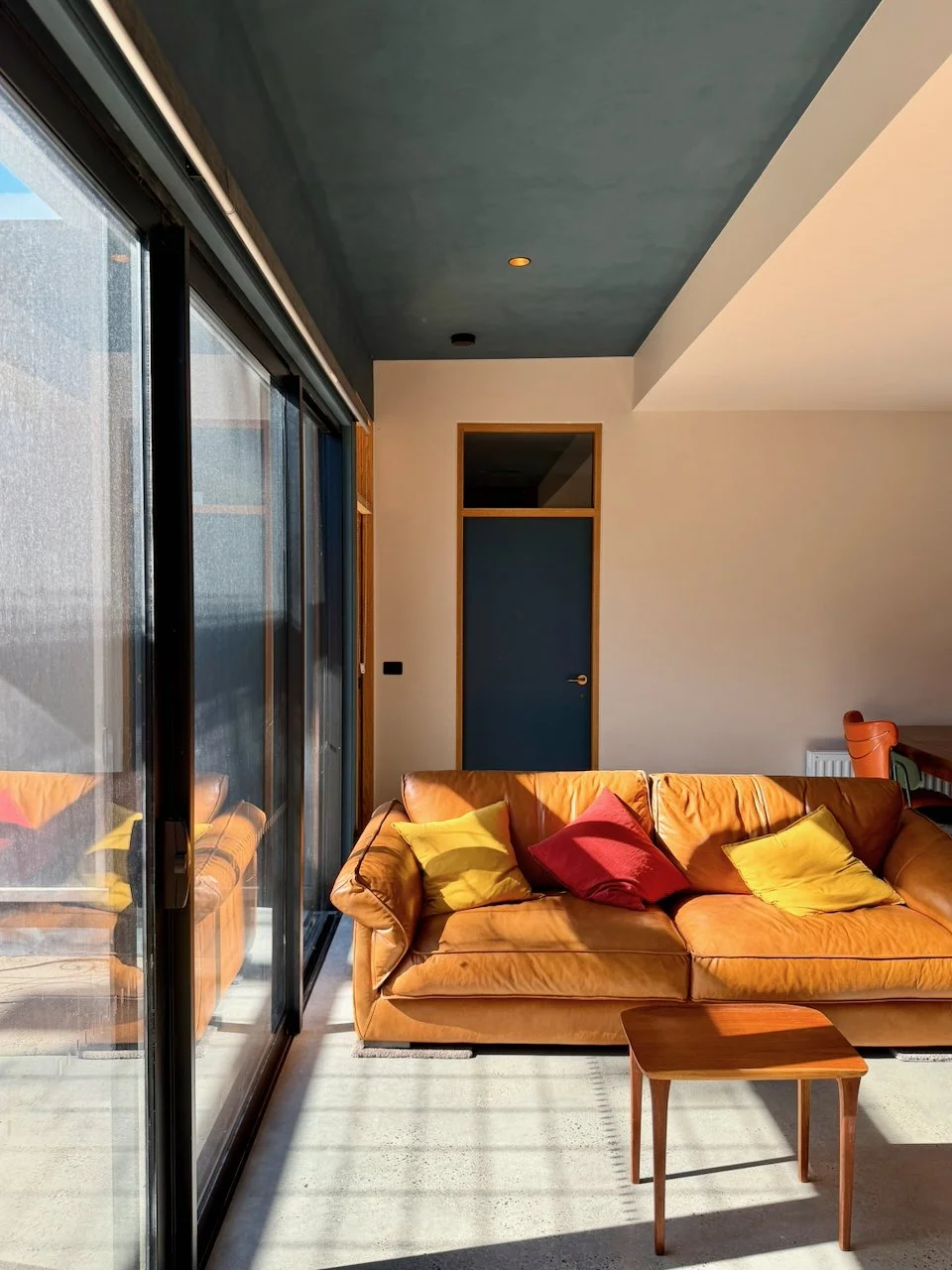 Living room with a large glass sliding door, a mustard-colored sofa with red and yellow pillows, a small wooden coffee table, and a door in the background.