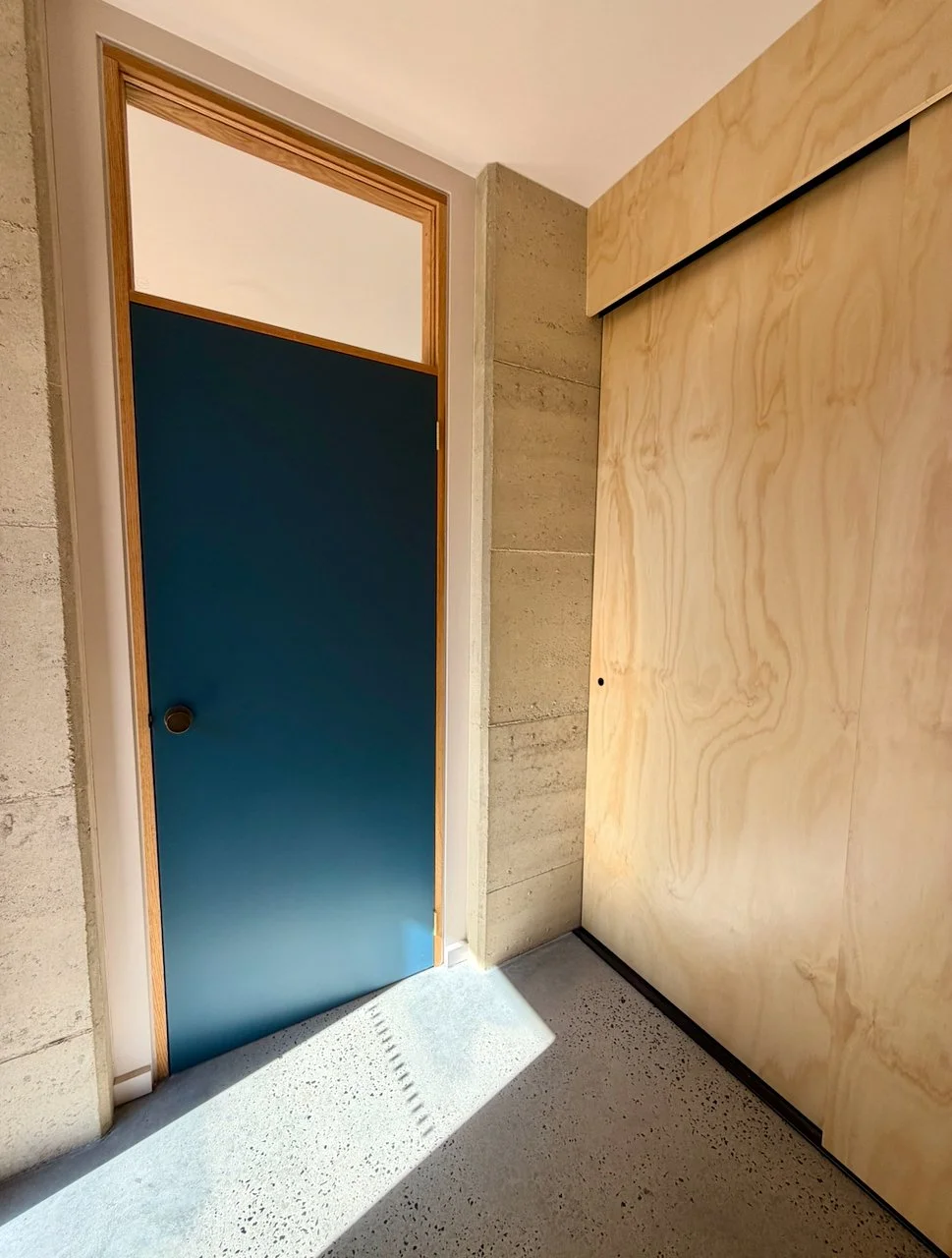 Interior corner of a room with a closed blue sliding door, a textured concrete wall to the left, a smooth light-colored wooden wall to the right, and a floor with popcorn texture. Sunlight casts shadows on the floor.