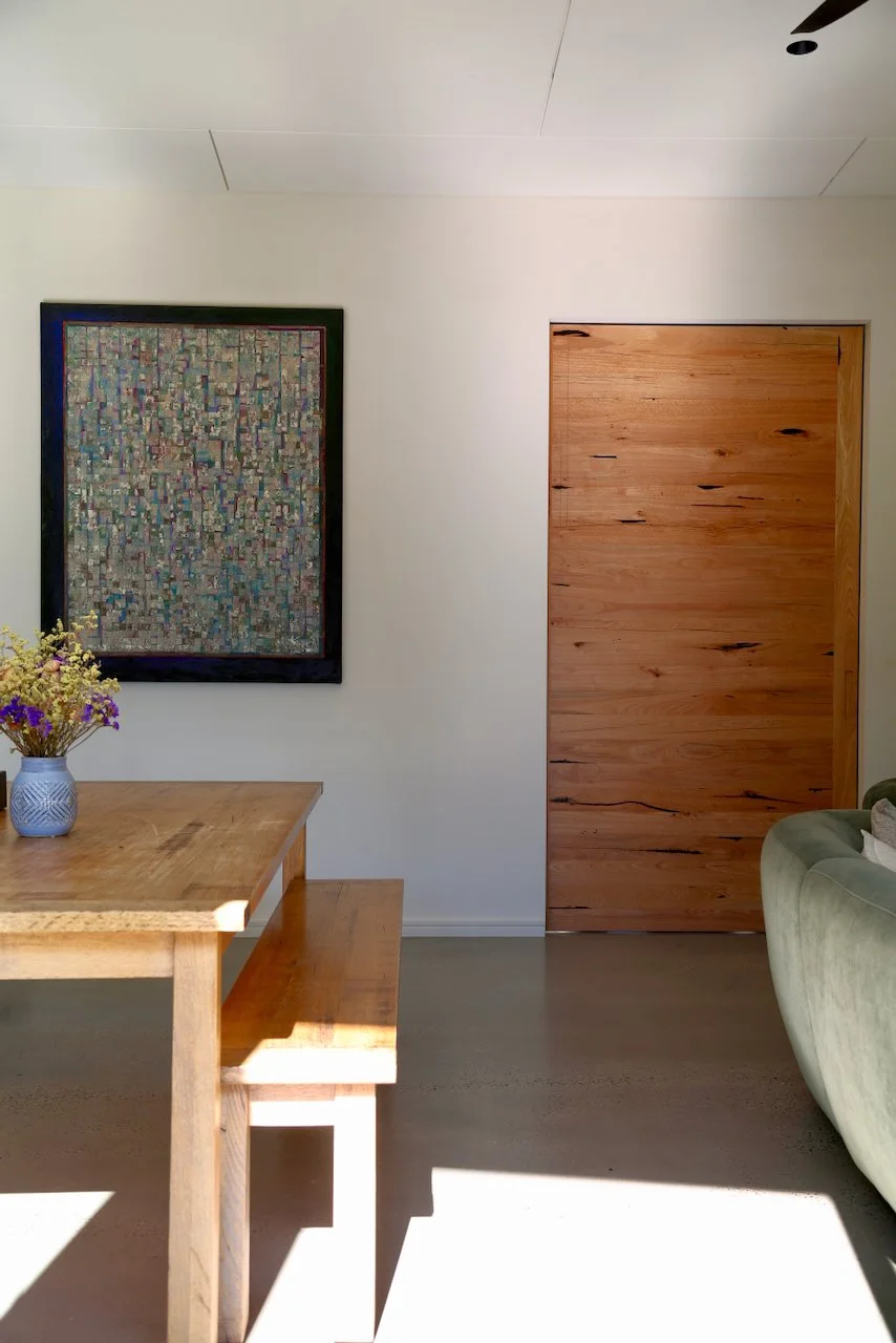 A corner of a room with a wooden door, a framed abstract artwork on the wall, a dining table with a vase of flowers, and part of a green sofa with a cushion, with sunlight casting shadows on the floor.