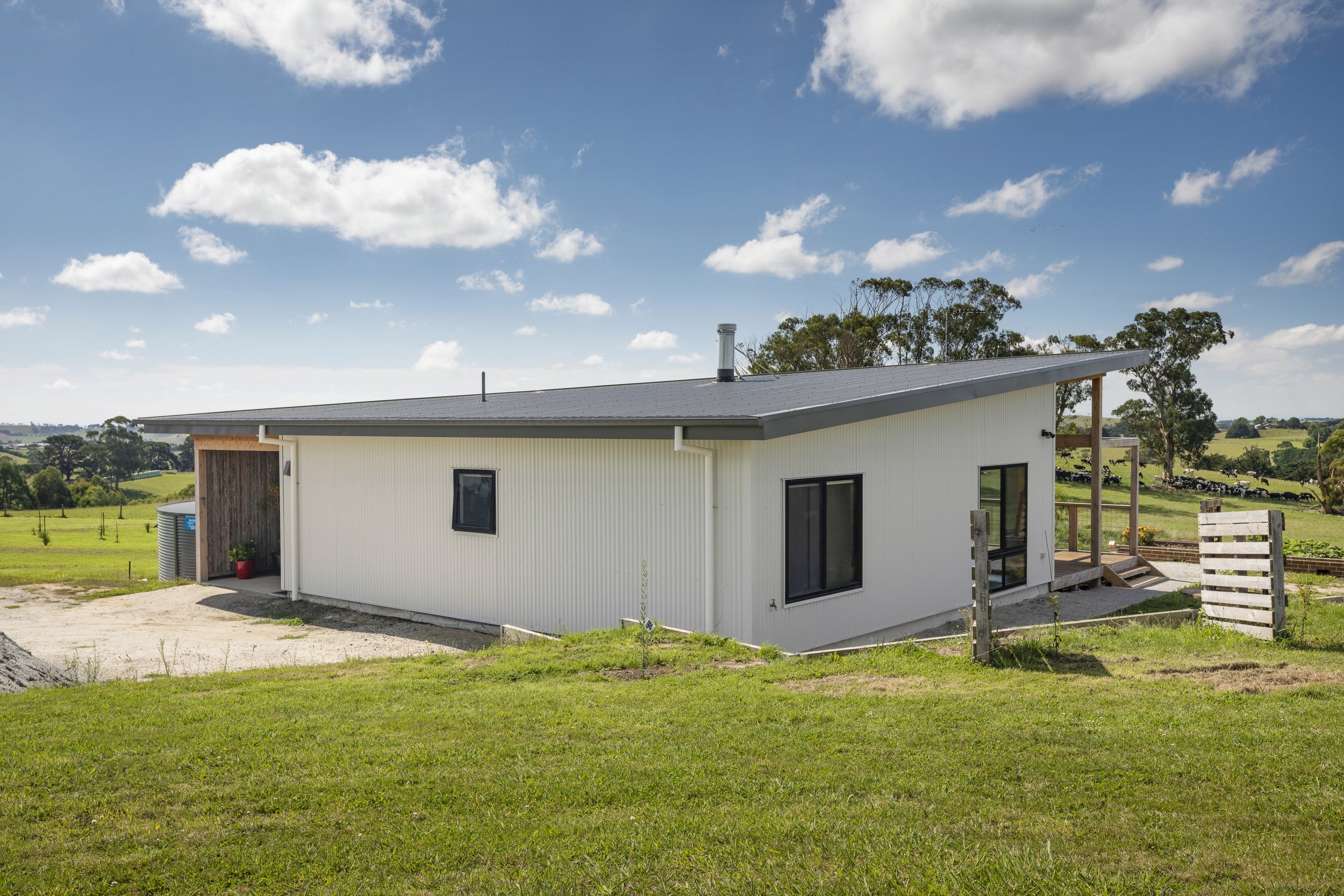 A modern white house with a sloped metal roof, situated in a rural landscape with green fields, trees, and a blue sky with scattered clouds.