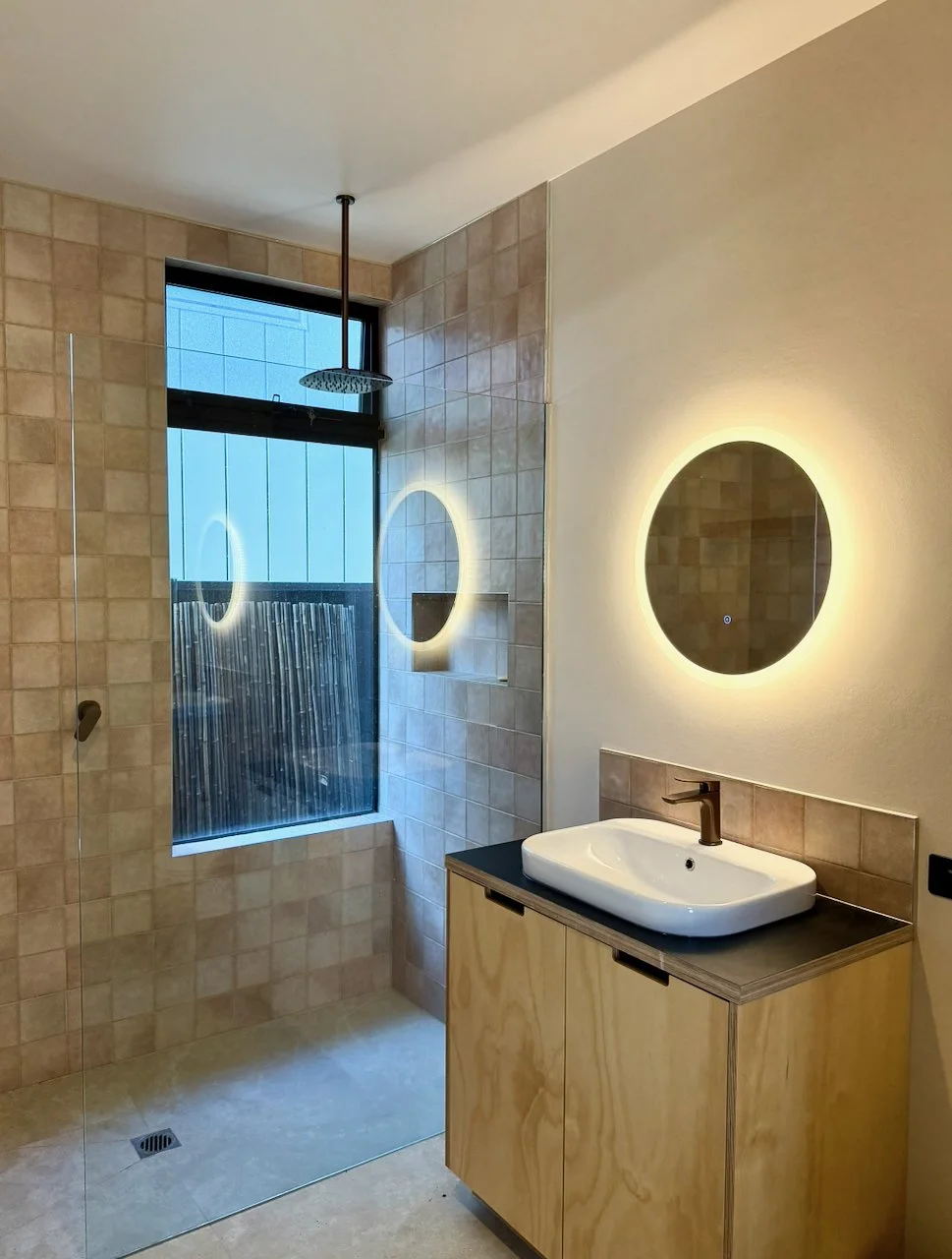 Modern bathroom with walk-in shower, round illuminated mirror, white oval vessel sink, wooden vanity, and a window behind the shower.