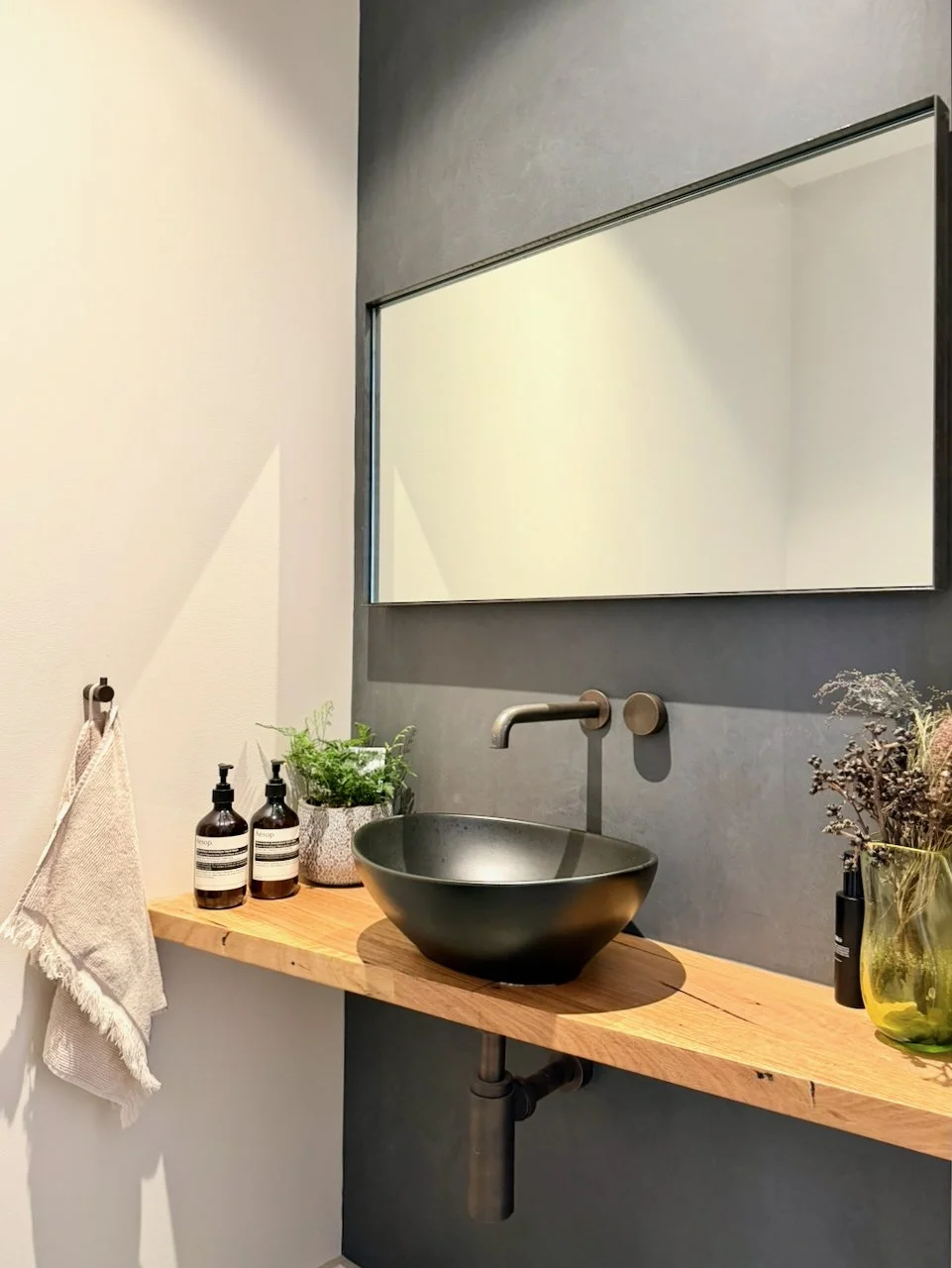 Bathroom vanity with a black vessel sink, wall-mounted faucet, large mirror, potted plant, soap dispensers, and dried flowers on a wooden countertop.