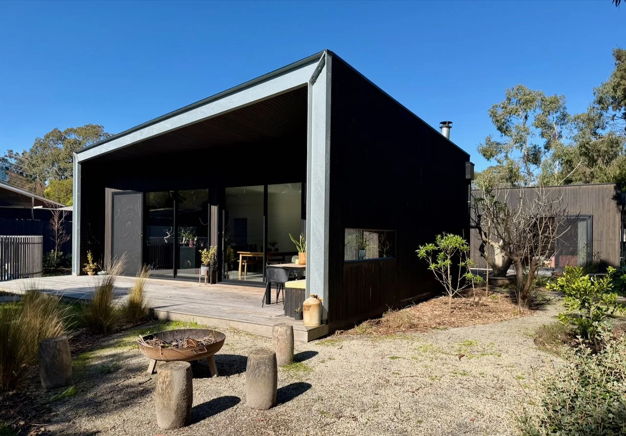 Modern black house with large glass sliding doors and a wooden deck, surrounded by landscaped yard with small trees, shrubs, and a gravel area with wooden stools and a fire pit, under a clear blue sky.
