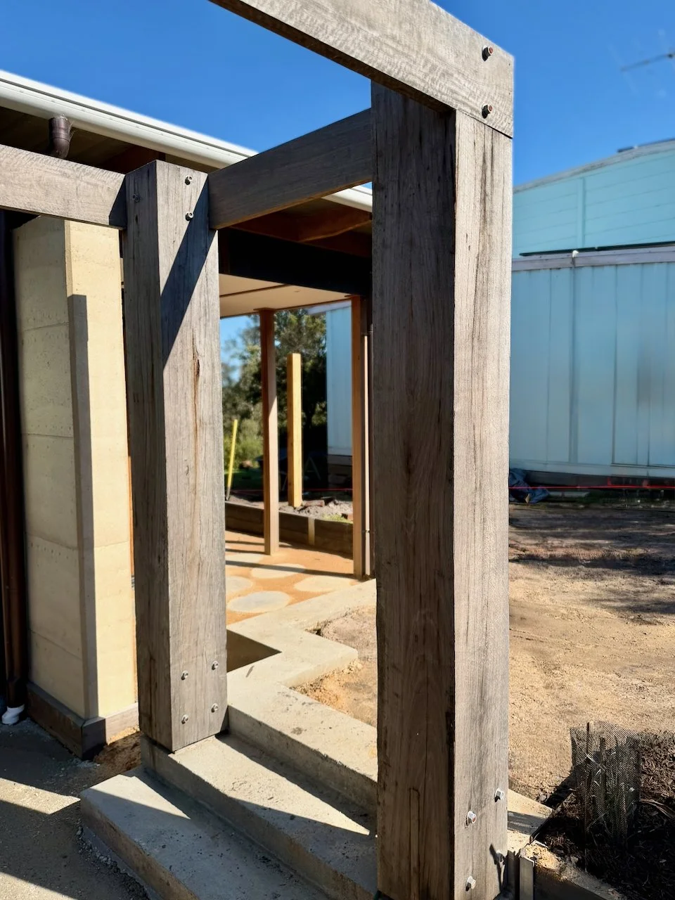 Wooden framing for a building under construction with concrete steps, between a beige wall and another building with white siding, on a clear blue sky day.