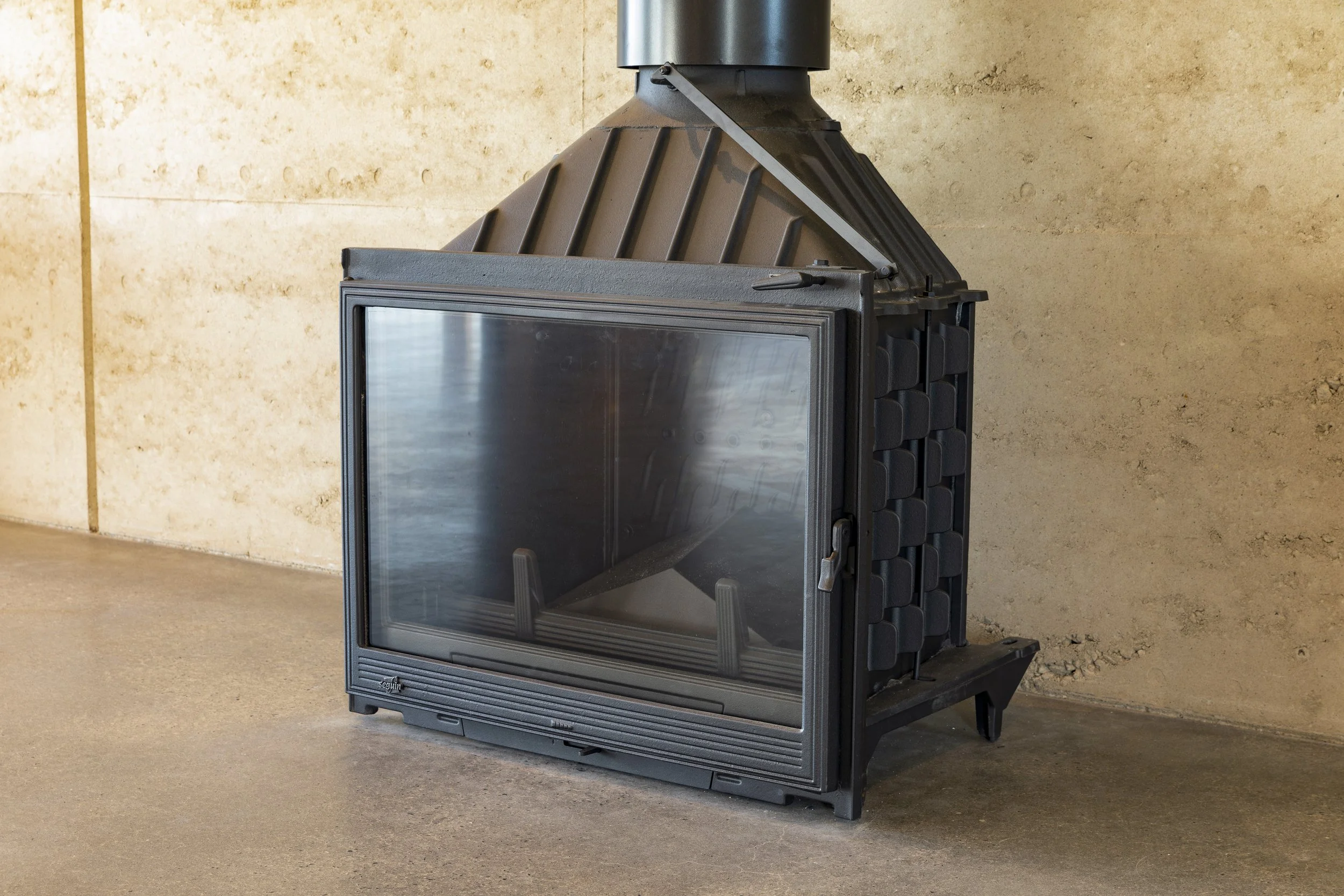 A black metal fireplace with a glass front and a sloped, ribbed hood, standing on a concrete floor against a textured beige wall.