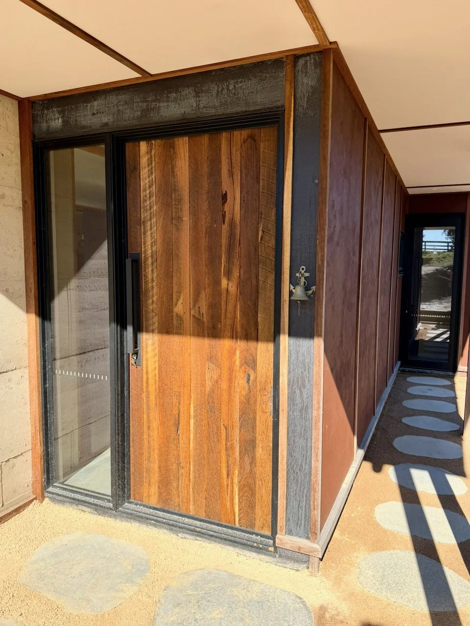 Exterior view of a building corner with a wooden sliding door, wood wall siding, and shadowed pathway with stepping stones.