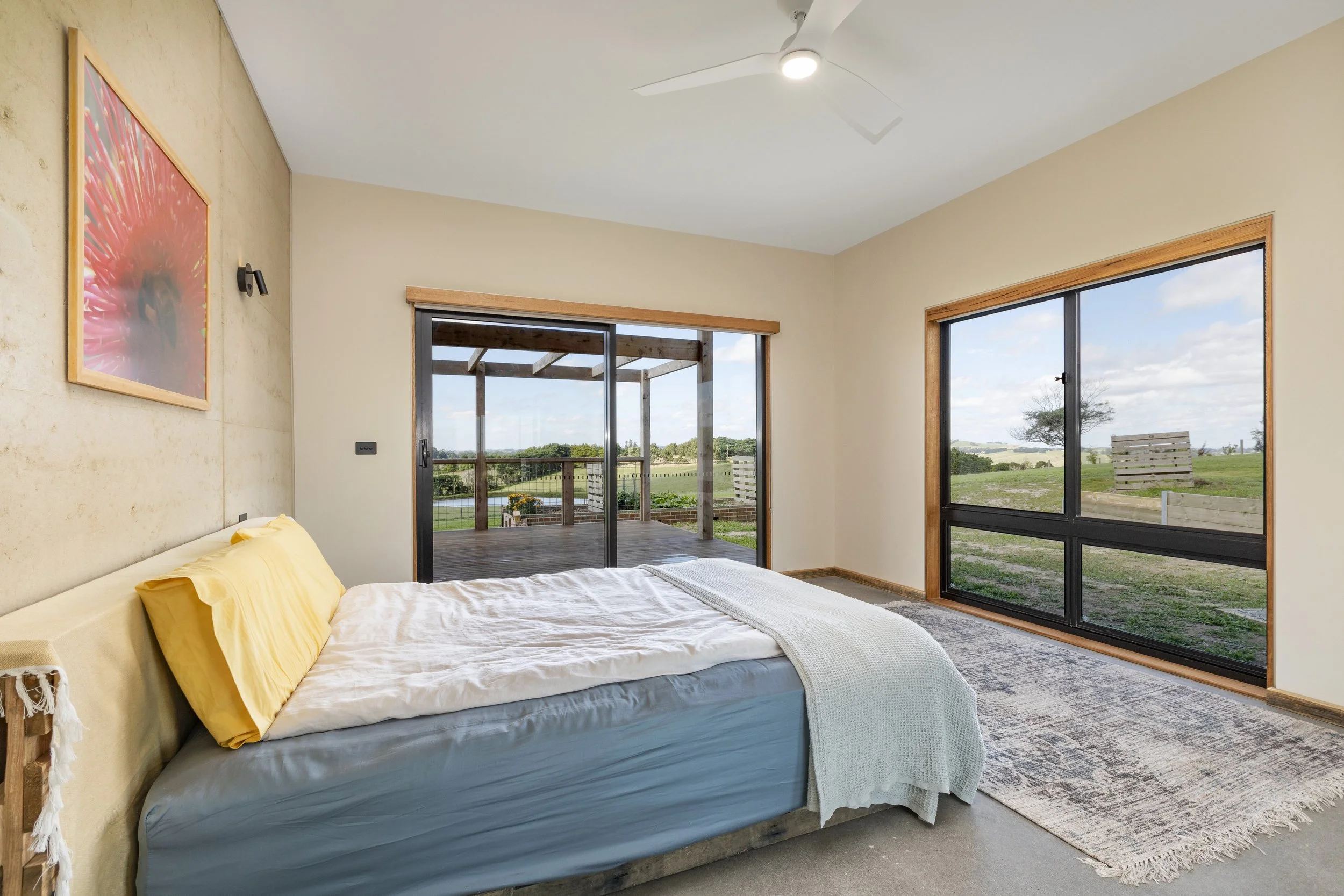 A bedroom with a bed featuring white and yellow bedding, large windows showing an outdoor deck and a rural landscape, and artwork on the beige wall.