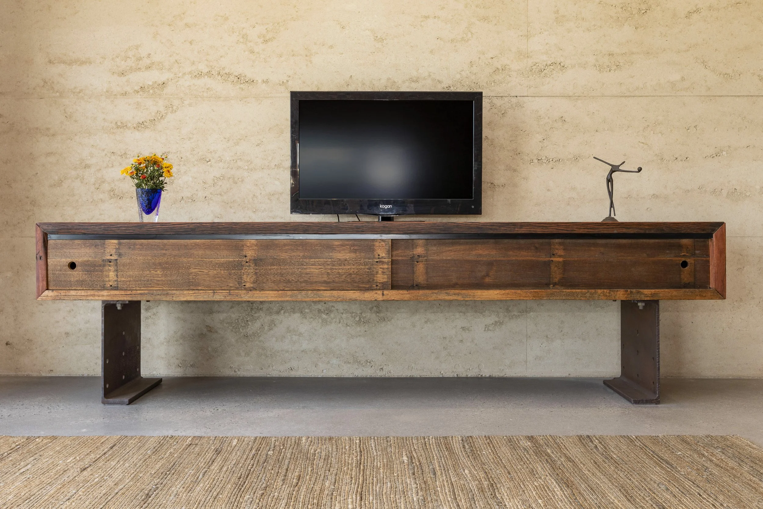 A wooden sideboard with a flat-screen television, a blue vase with yellow flowers, and a metal sculpture on top, against a beige wall with a textured surface.