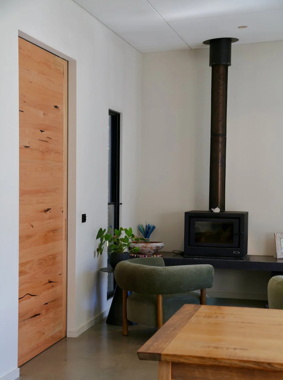 Living room corner with a wood stove pipe, green armchair, small side table with plant, and wooden door.
