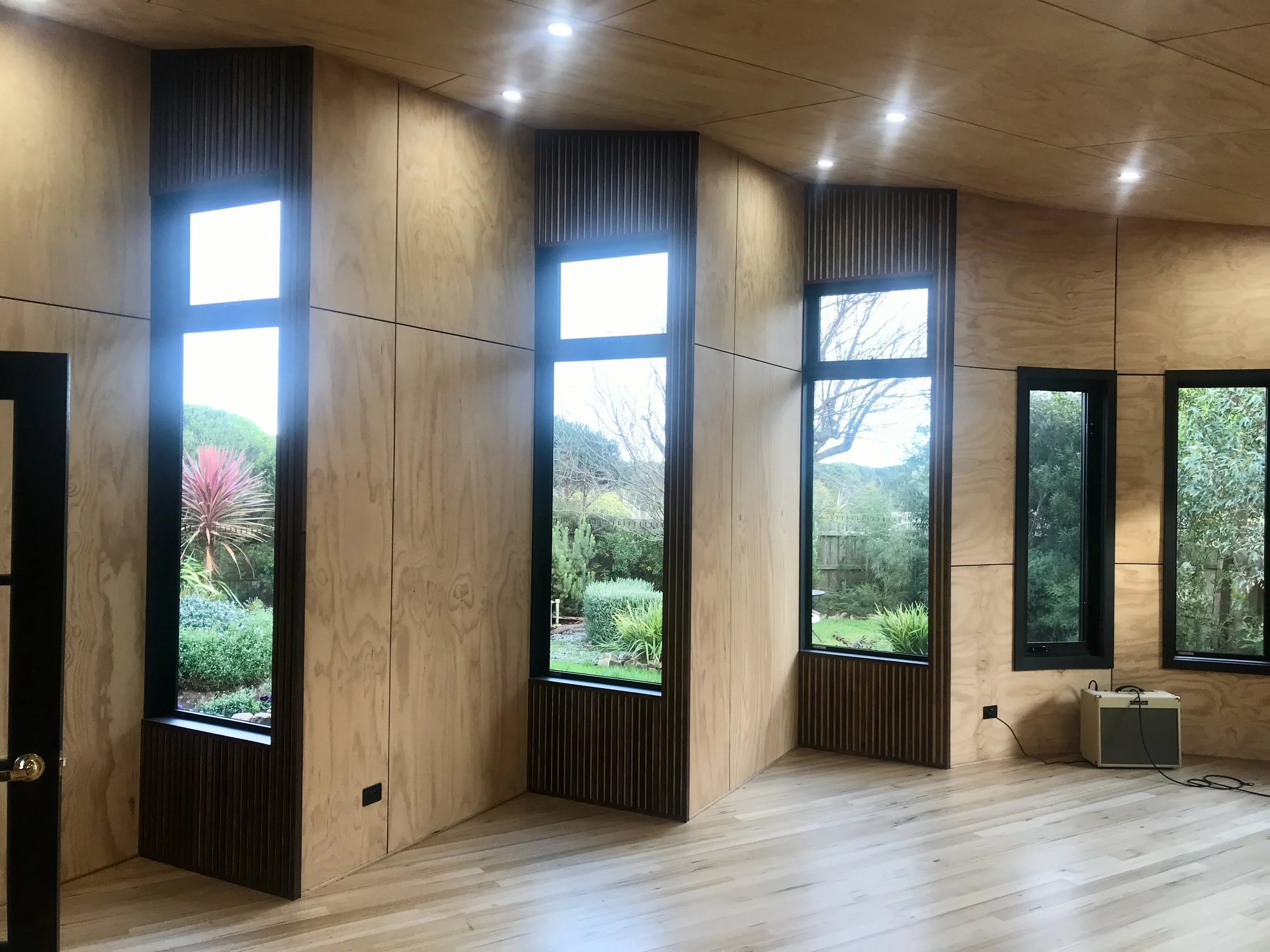 Interior of a room with light-colored wood panel walls and ceiling, three tall windows with black trim showing a garden outside, and a small white heater plugged into an electrical outlet on the right side.