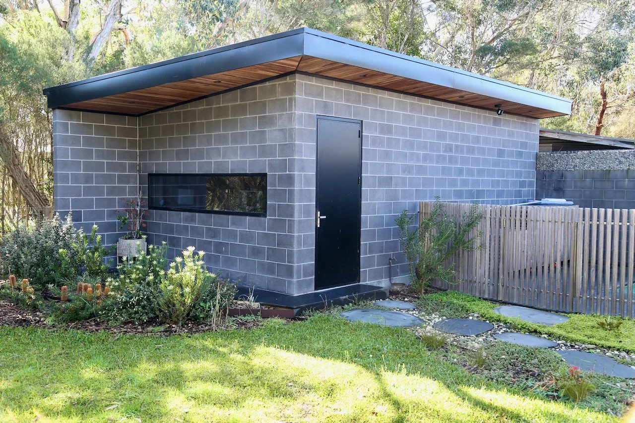Small modern building with a black door, a horizontal window, and a sloped roof, surrounded by a garden with various plants and a stone pathway.