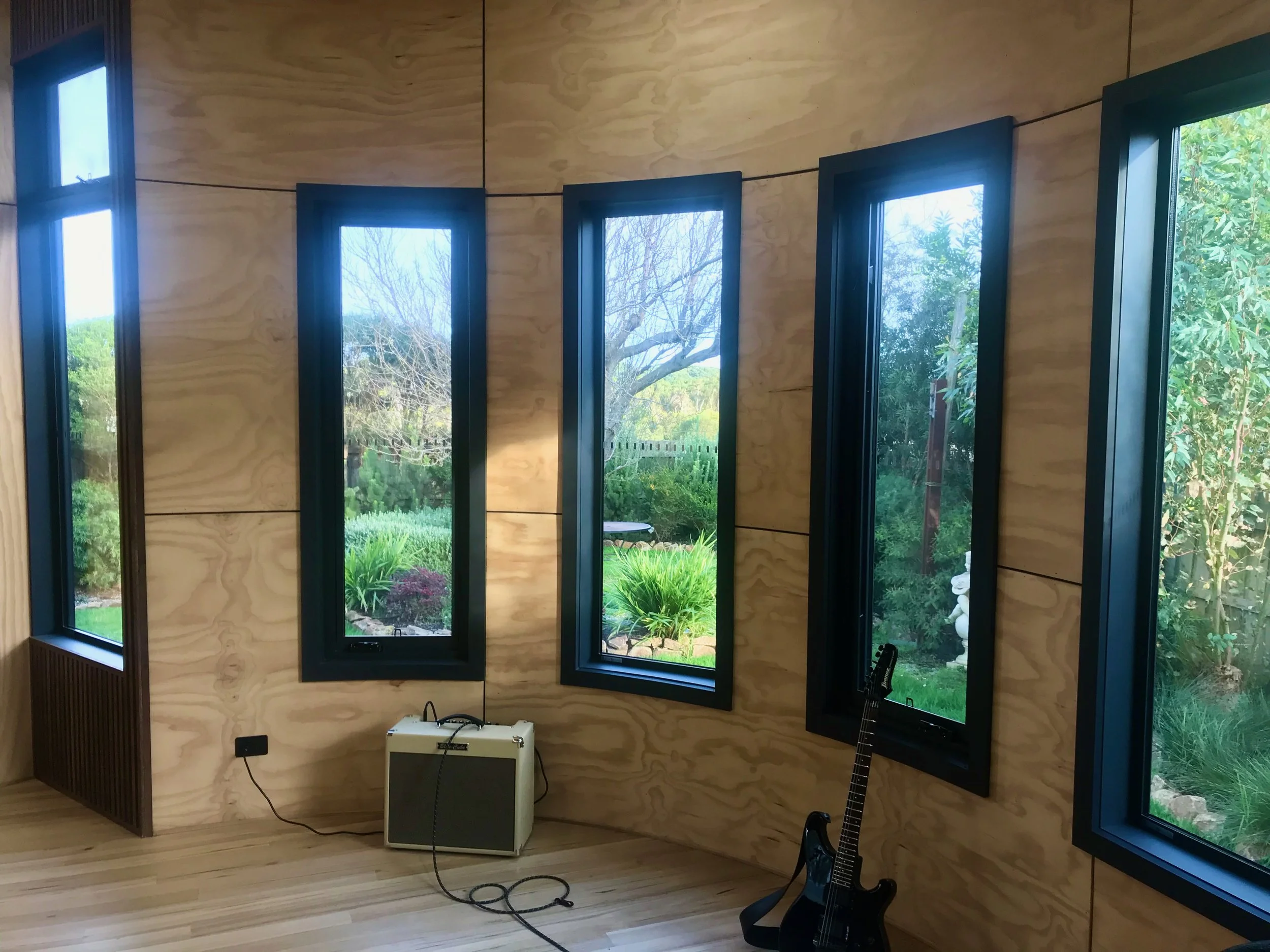 Room with large windows showing a garden outside, wooden walls and floor, a white amplifier on the floor, a black guitar on a stand, and an electric guitar leaning against the wall.