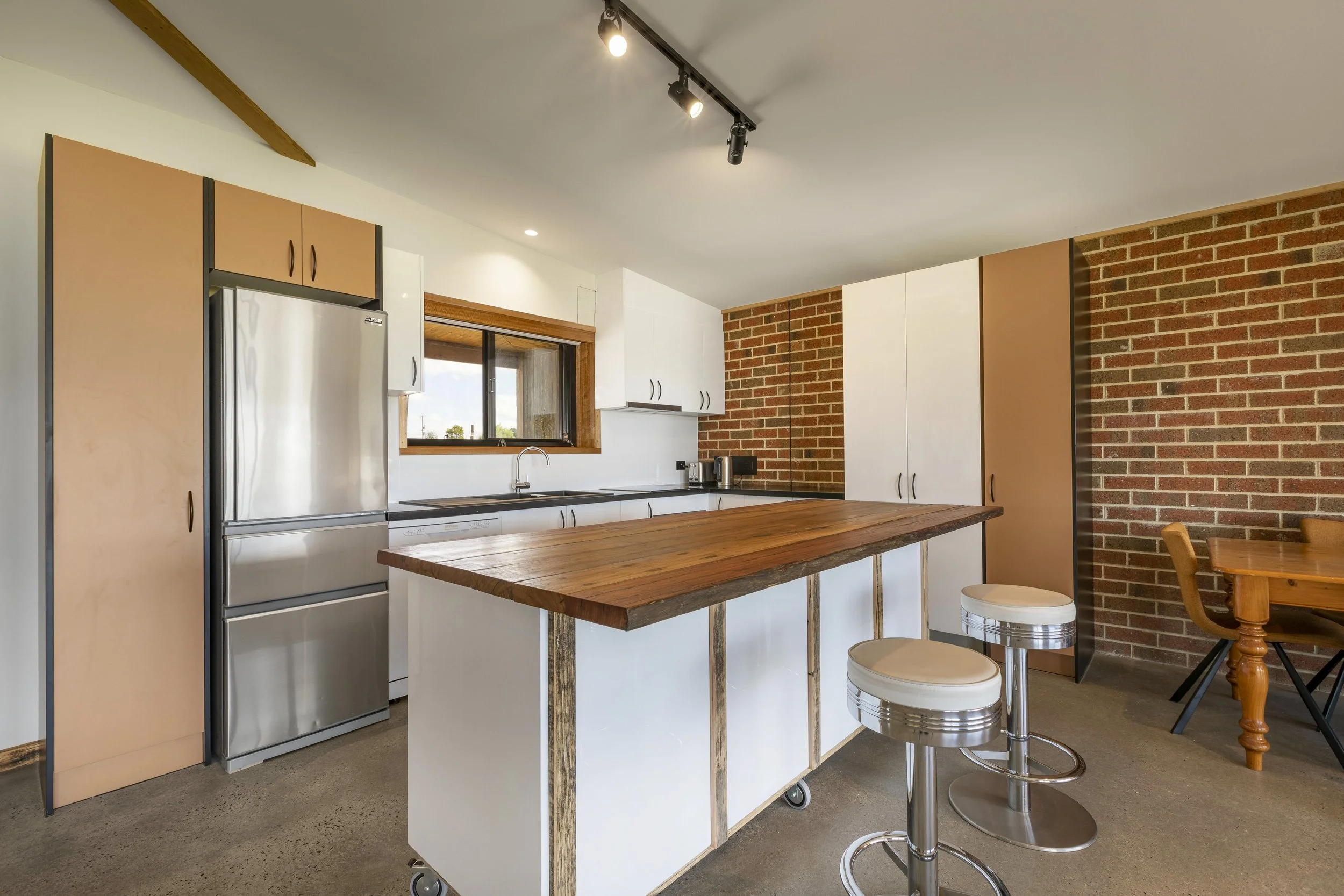 Modern kitchen with white and wood cabinetry, a brick accent wall, a stainless steel refrigerator, a window above the sink, and a wooden kitchen island with metal wheels. There are two bar stools and a dining area with a wooden table and chairs.