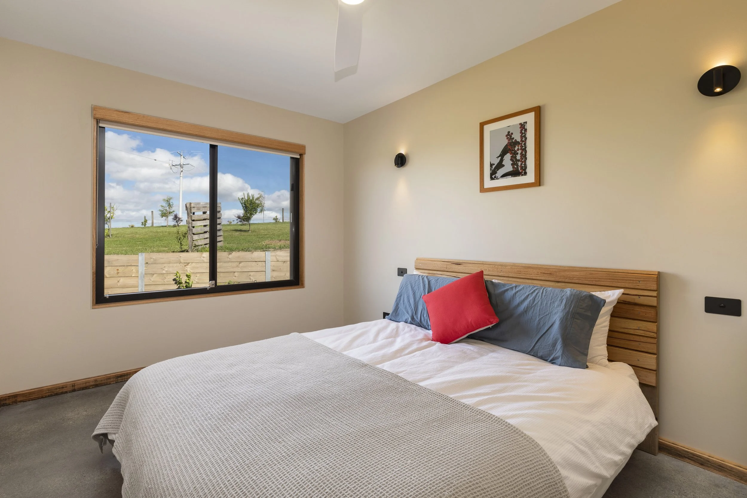 A bedroom with a large bed, blue and red pillows, and a view of a backyard through a sliding glass door. The room has beige walls, a wooden headboard, and modern wall-mounted lights.