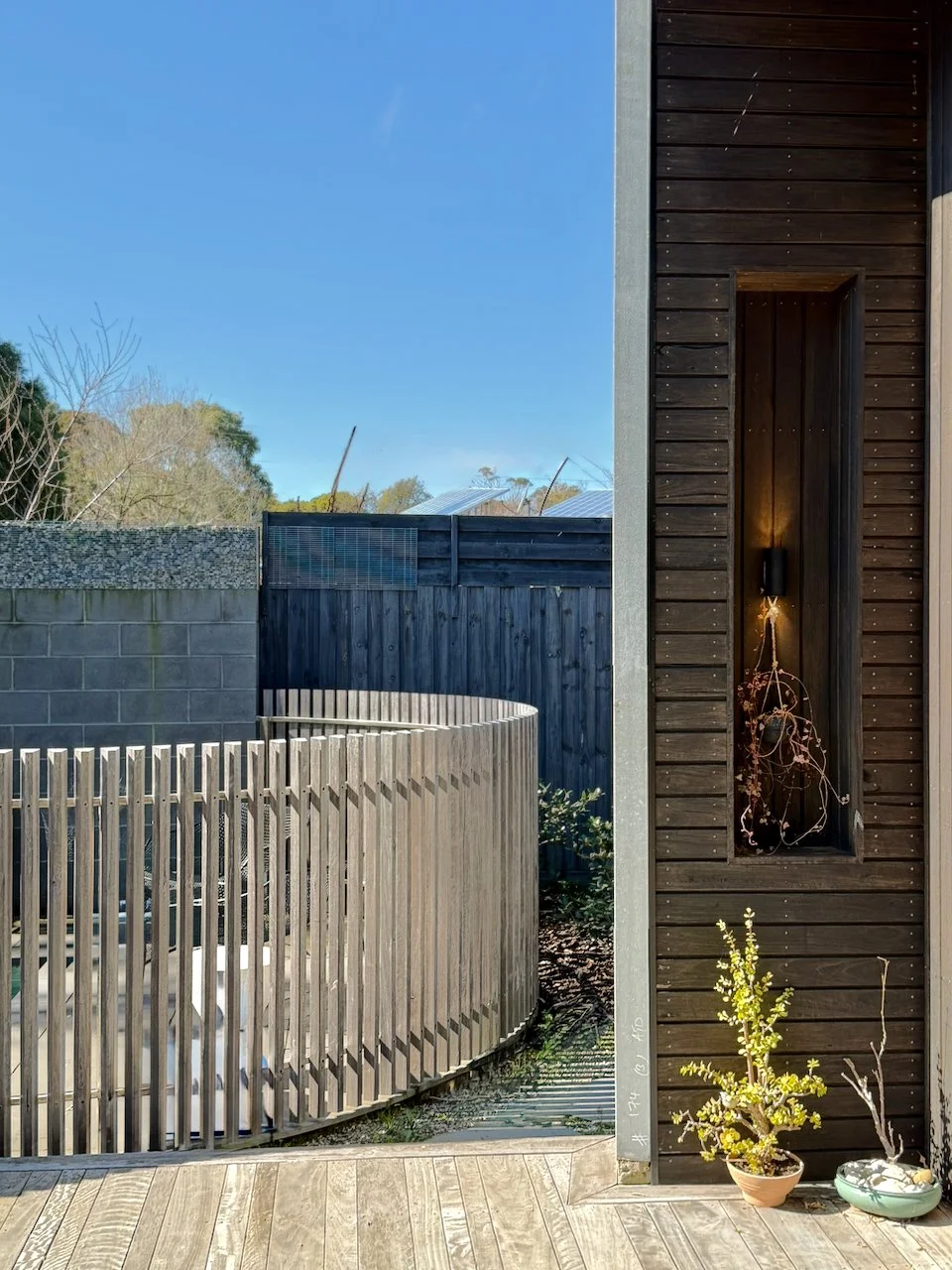 Part of a modern exterior with a wooden deck, a dark wooden wall with a niche holding a plant, and a plant in a pot on the floor, with a rounded wooden fence and neighboring structures visible in the background.