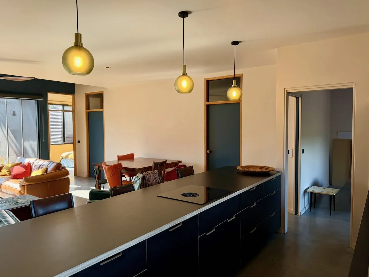 Open-concept living space with a kitchen island, three hanging pendant lights, dining table with chairs, and a living area with a sofa near a large window.