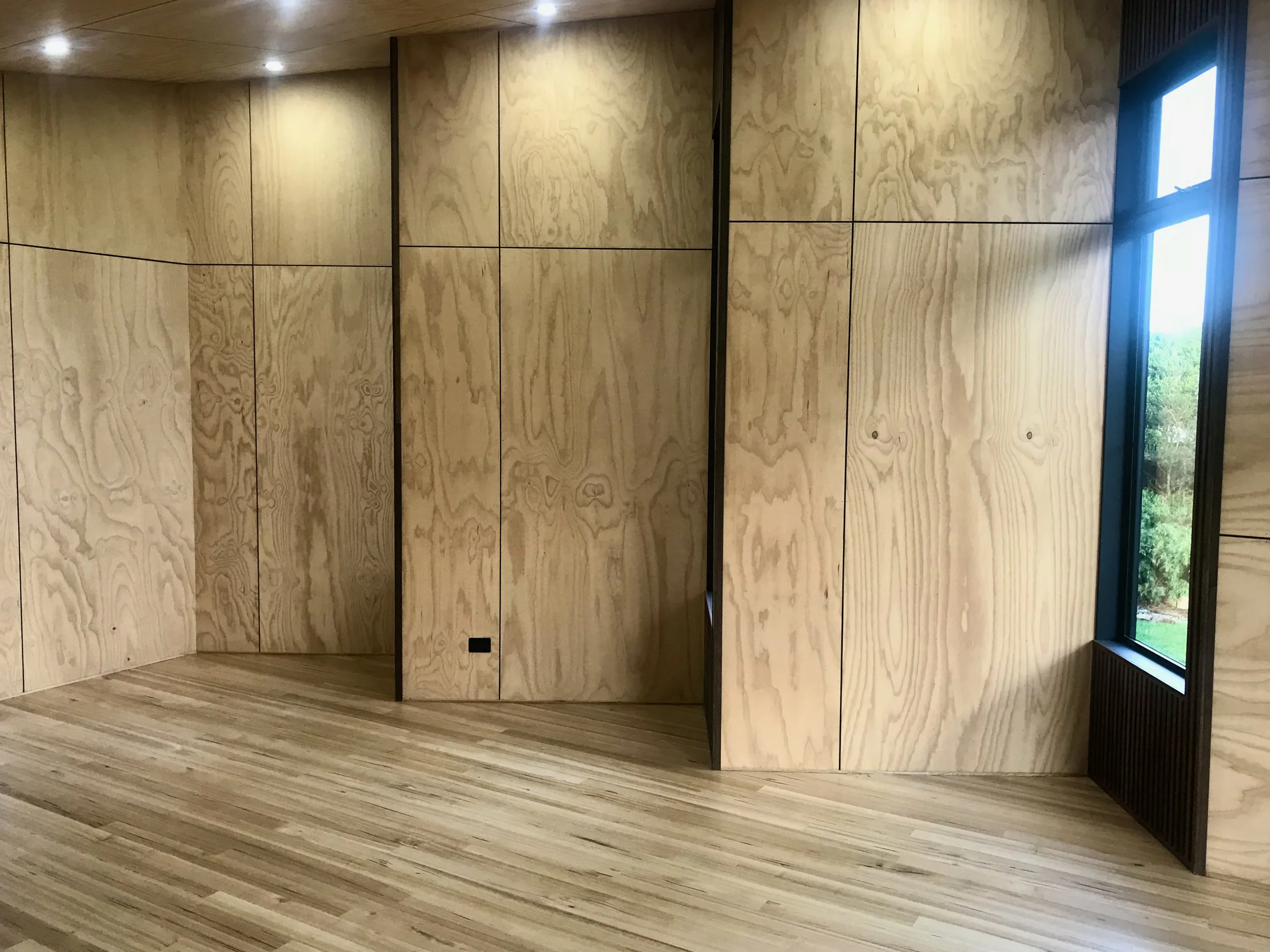 Interior space with light-colored wooden walls and flooring, a corner of a black framed window, and ceiling lights.