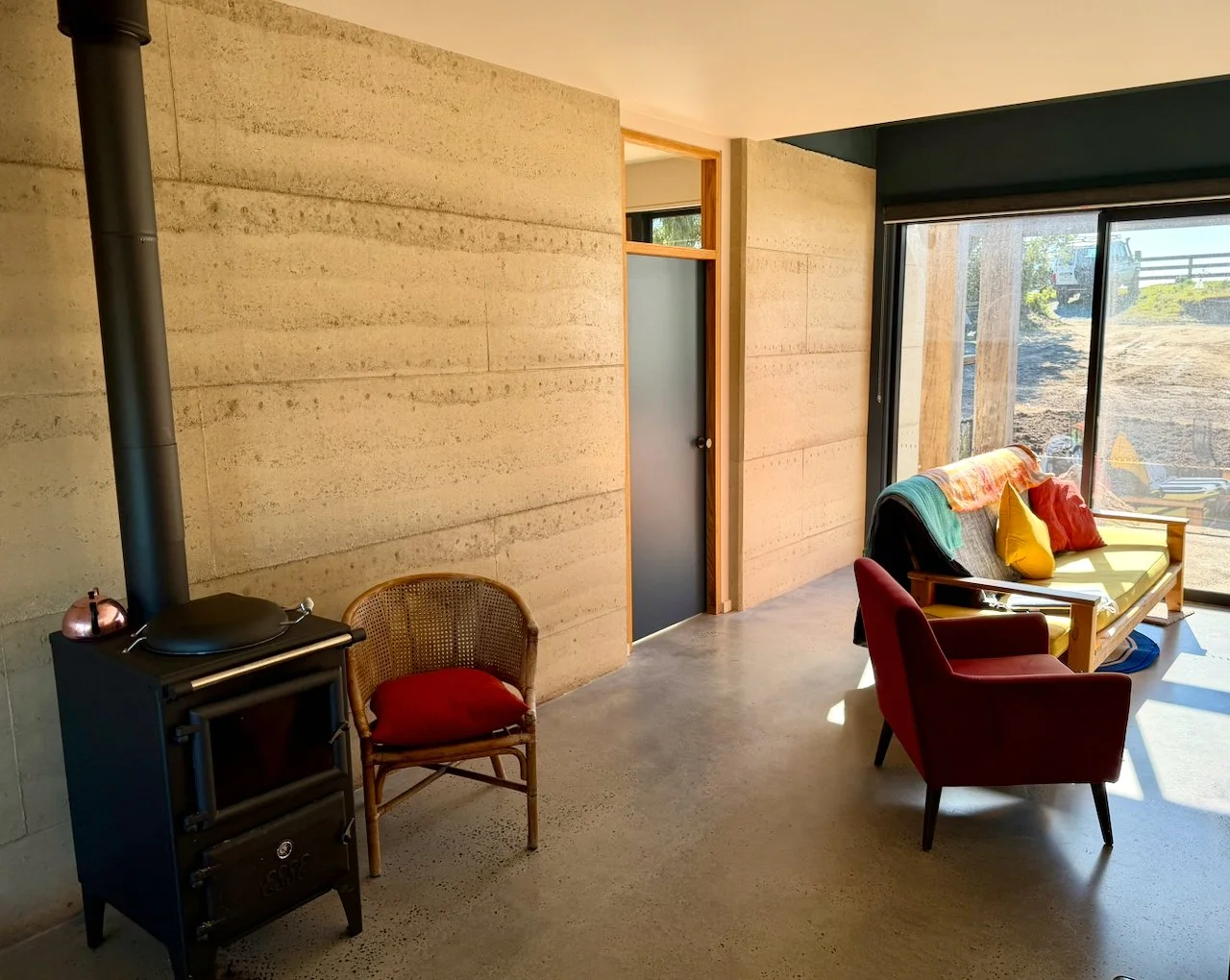 Interior of a modern living room with a concrete floor, beige stone walls, a black wood stove with a copper kettle on top, a rattan chair with a red cushion, a red armchair, and a wooden bench with yellow, red, and green pillows near large sliding gl