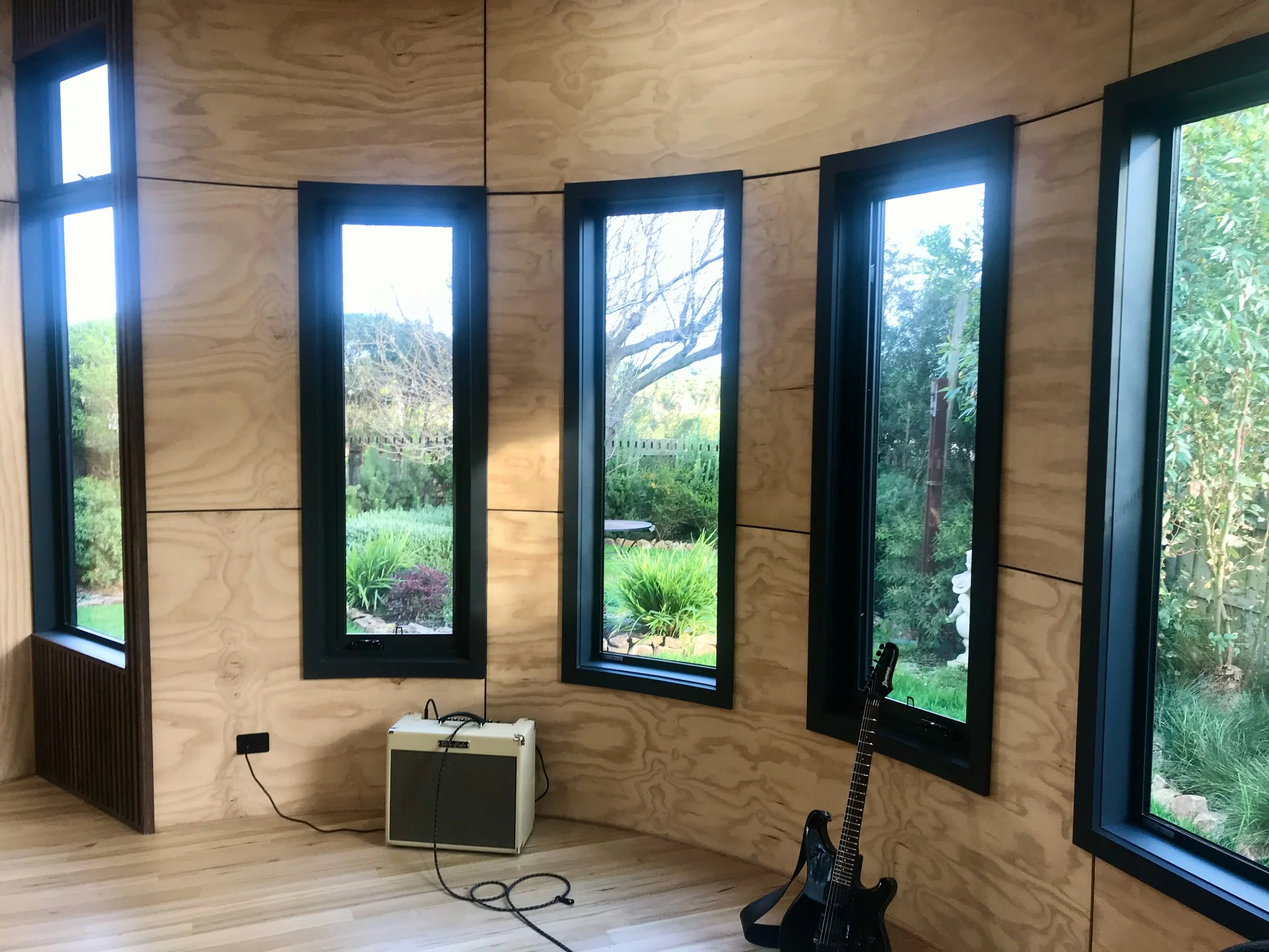 Interior of a room with wooden wall paneling and large black-framed windows showing an outdoor garden with trees and greenery. There is a black electric guitar standing on a stand, an amplifier, and a guitar cable on the floor.