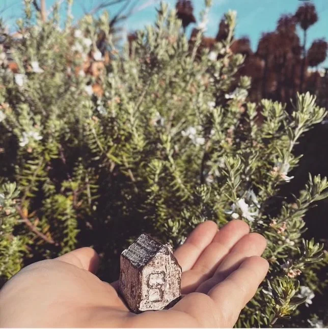 A hand holds a small rock that looks like a miniature houses with gray and white colors, in front of green shrubbery with small white flowers and blurred trees in the background.