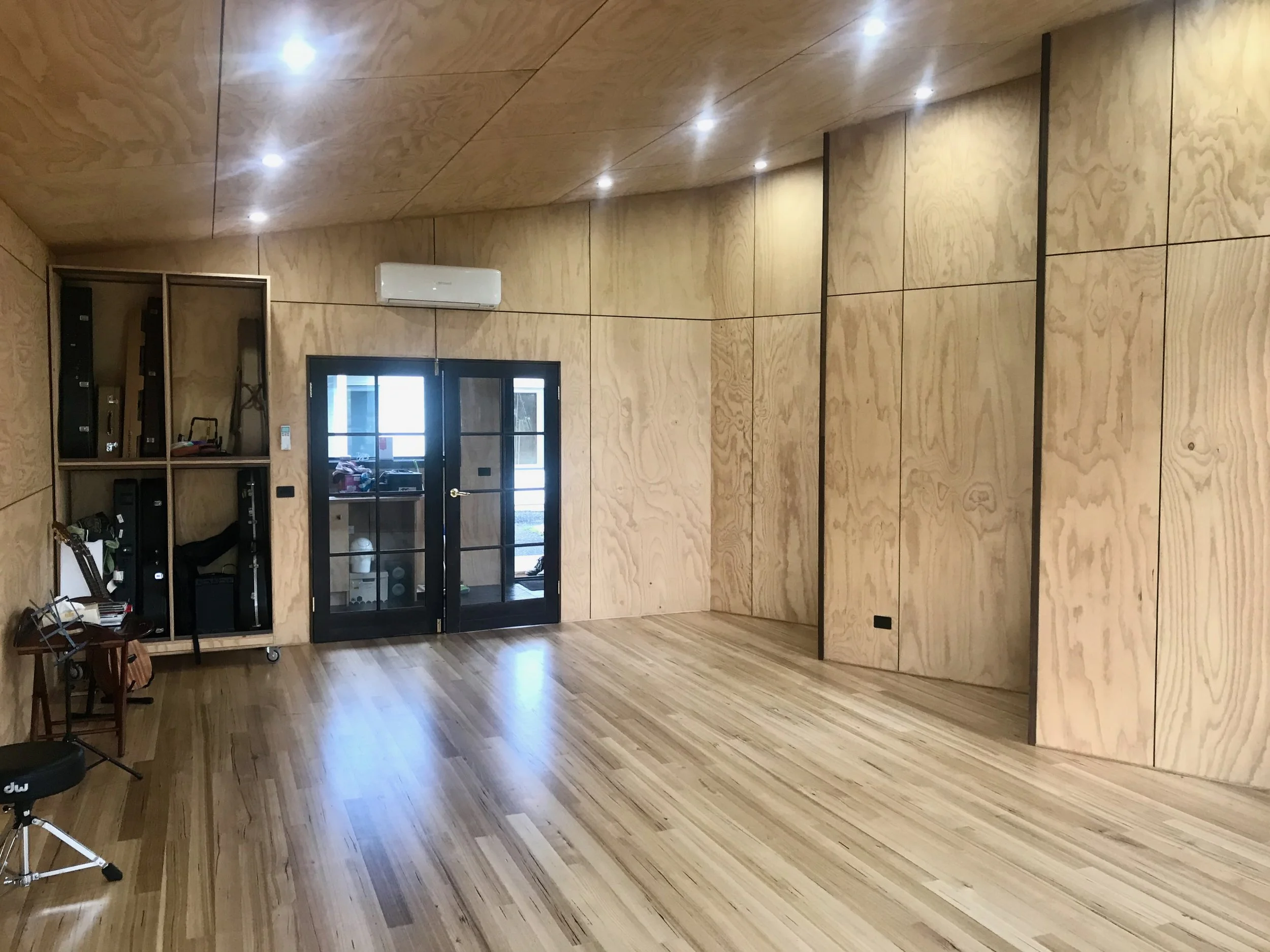 Empty room with wooden floors, wood-paneled walls, ceiling lights, a black-framed glass door, and some furniture including a cabinet and chair.