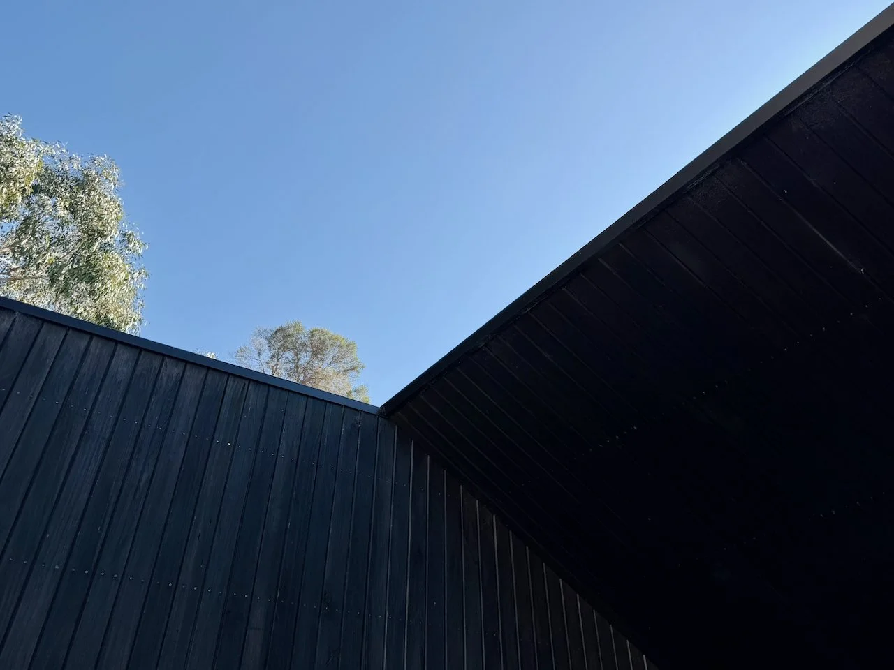 Close-up of the dark-colored roof and walls of a modern building with a clear blue sky and some trees in the background.