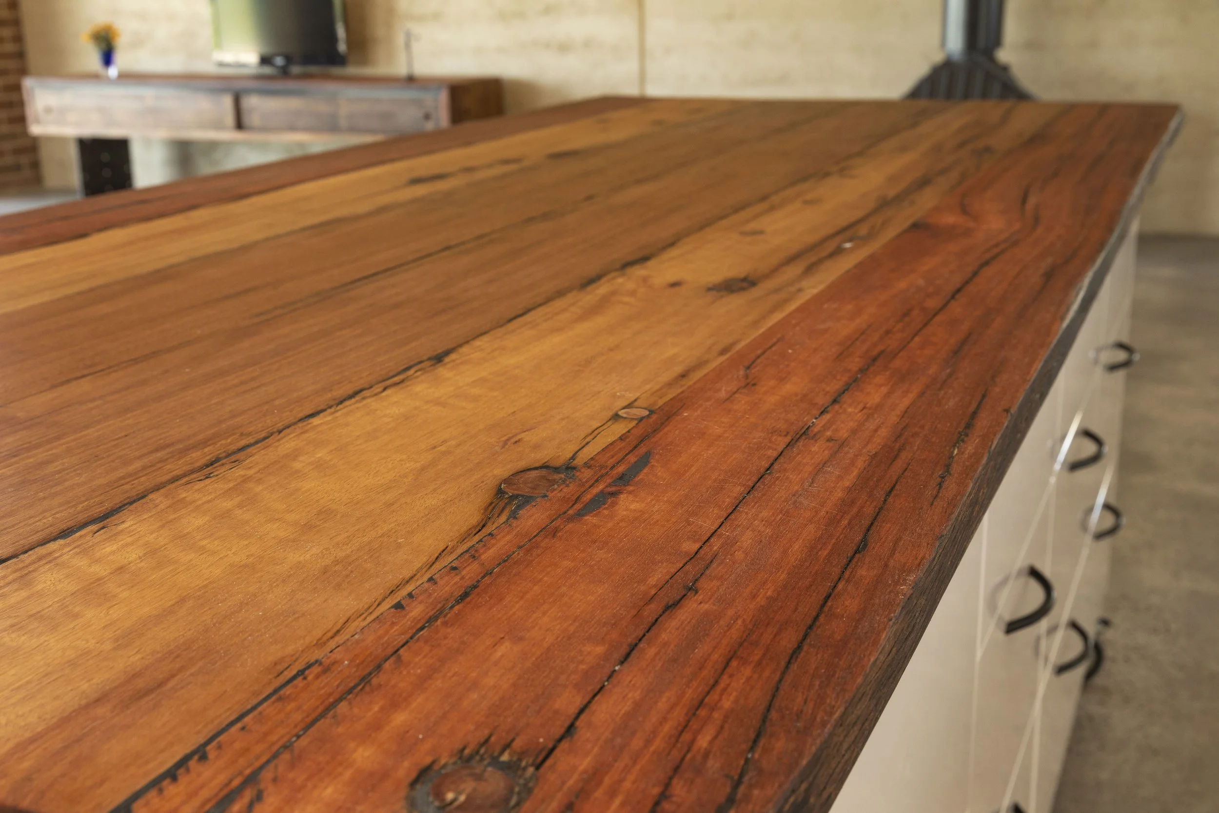 Close-up view of a polished wooden tabletop with natural wood grain and knots, situated in a room with a brick wall, a TV, and a wooden cabinet in the background.