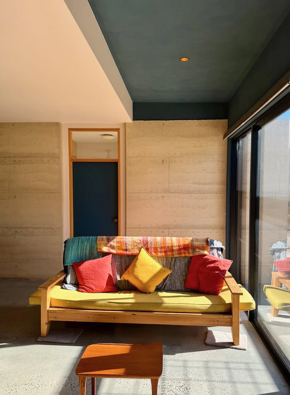 Sunlit living room with a wooden sofa, yellow cushions, red and green throw blankets, and large glass sliding doors.