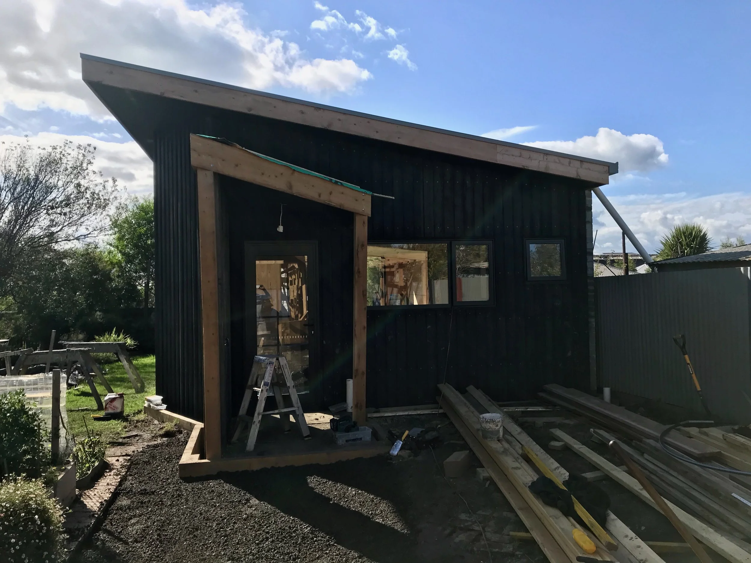 Construction site with a partially built black building, wooden frames, ladders, and tools, with a blue sky and some clouds in the background.