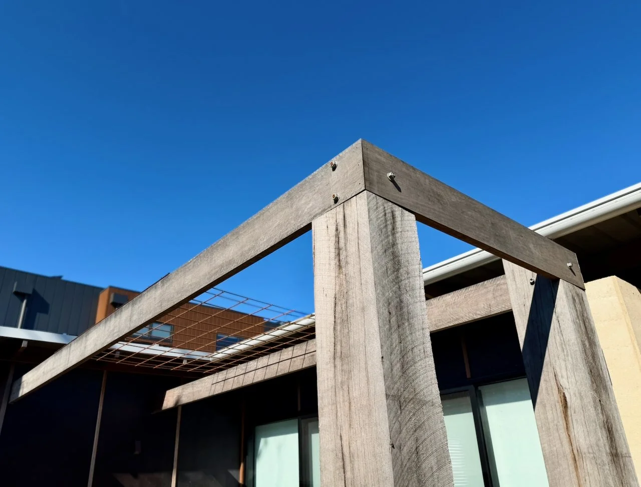 Part of a modern building under construction with wooden beams, a metal grid, and large glass windows, against a clear blue sky.