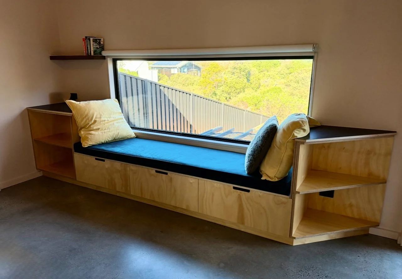 Wooden window seat with storage drawers, pillows, and a black cushion, situated below a large rectangular window showing an outdoor view of trees, a metal railing, and rooftops.