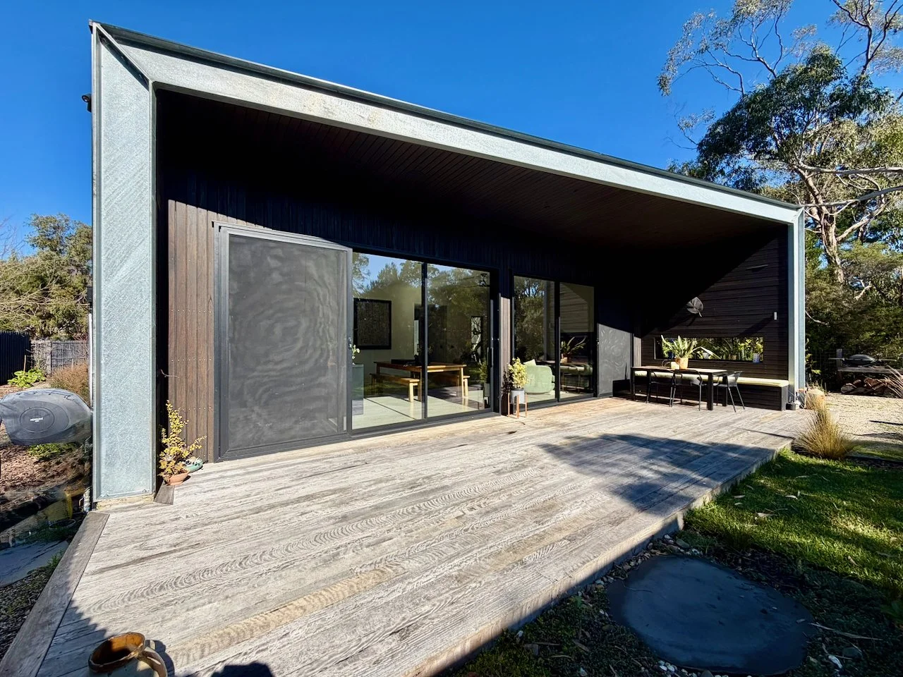 Modern house with large sliding glass doors, a wooden deck, outdoor dining area, and surrounding trees under a clear blue sky.