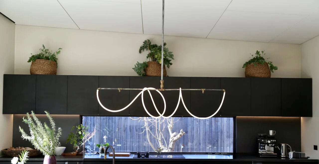 Modern kitchen with black cabinetry, a large window overlooking a backyard with trees, and decorative plants in woven baskets on top of the cabinets. An artistic white light fixture hangs from the ceiling.