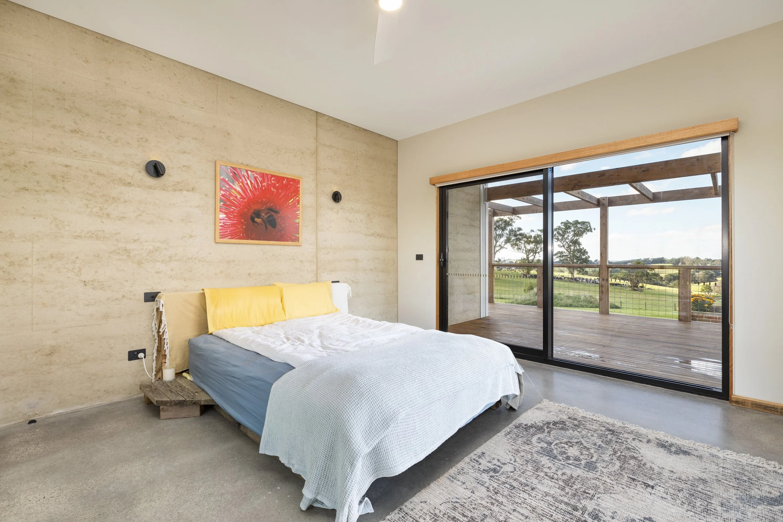 A bedroom with beige textured wall, a bed with yellow pillows and light-colored bedding, a framed flower art on the wall, and large sliding glass doors leading to a wooden balcony with a view of green trees and a sunny sky.