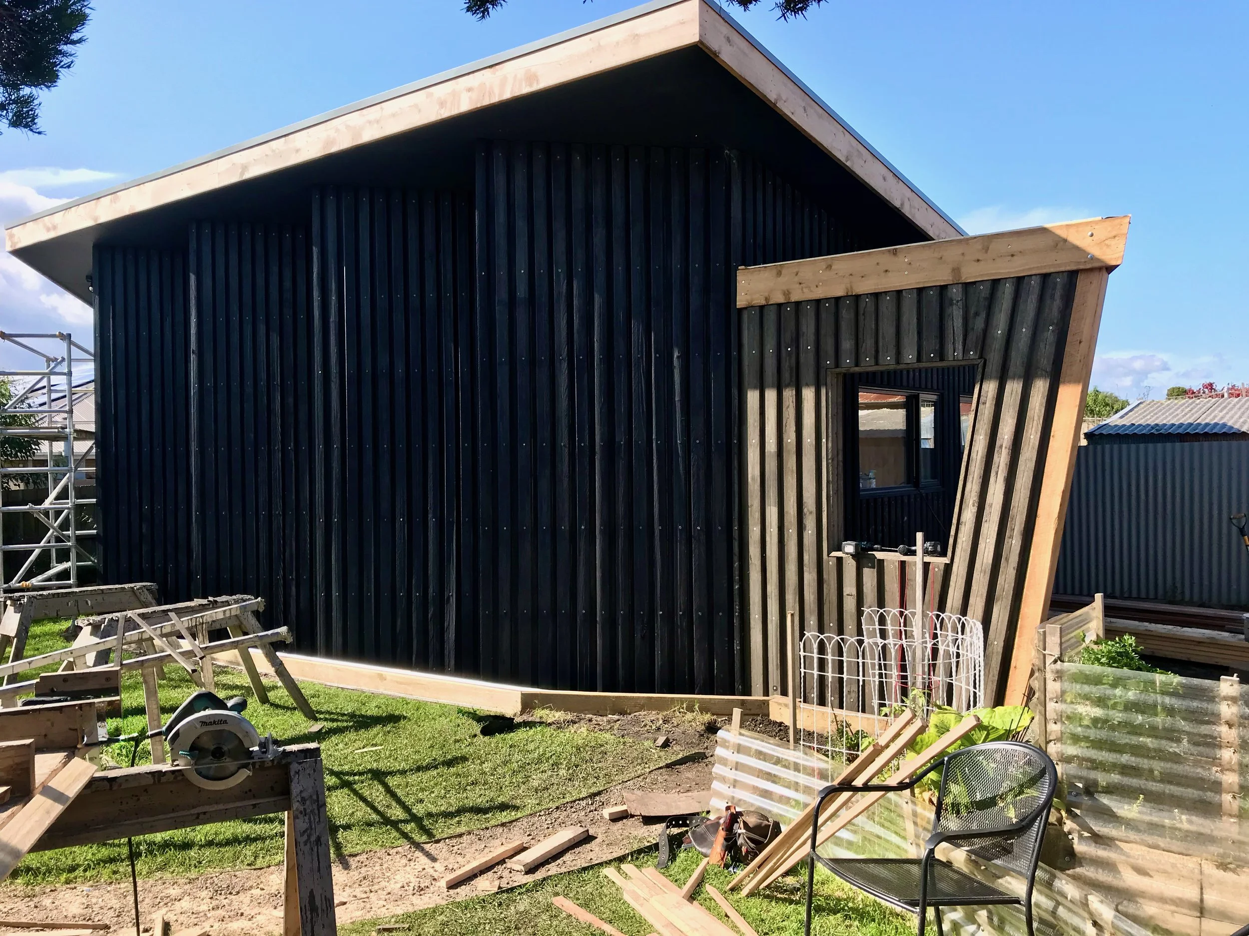 A house under construction with black vertical siding and unfinished wooden trim, surrounded by construction tools and materials in a backyard.