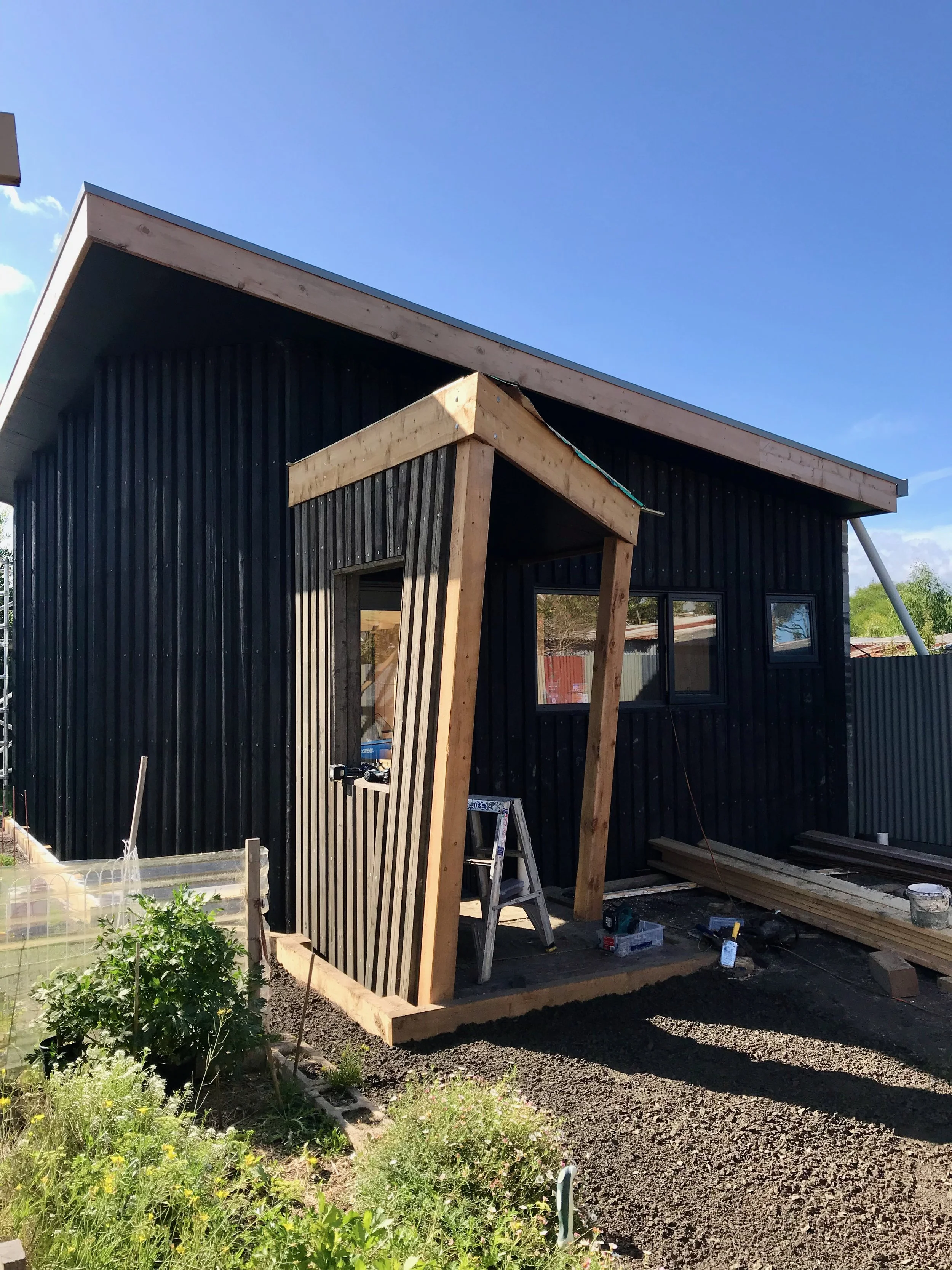 Construction site of a small building with black vertical siding and wooden framing on the entrance, with garden plants and a vegetable bed in the foreground, and a clear blue sky overhead.