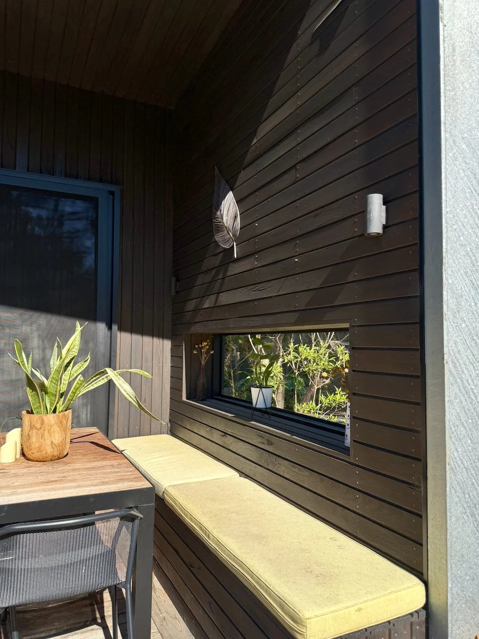 A cozy outdoor patio corner with a long built-in seating area with pale yellow cushions, a table with a potted plant, a chair, a narrow horizontal window with plants outside, a dark wooden wall, a decorative feather, and a wall-mounted light fixture.