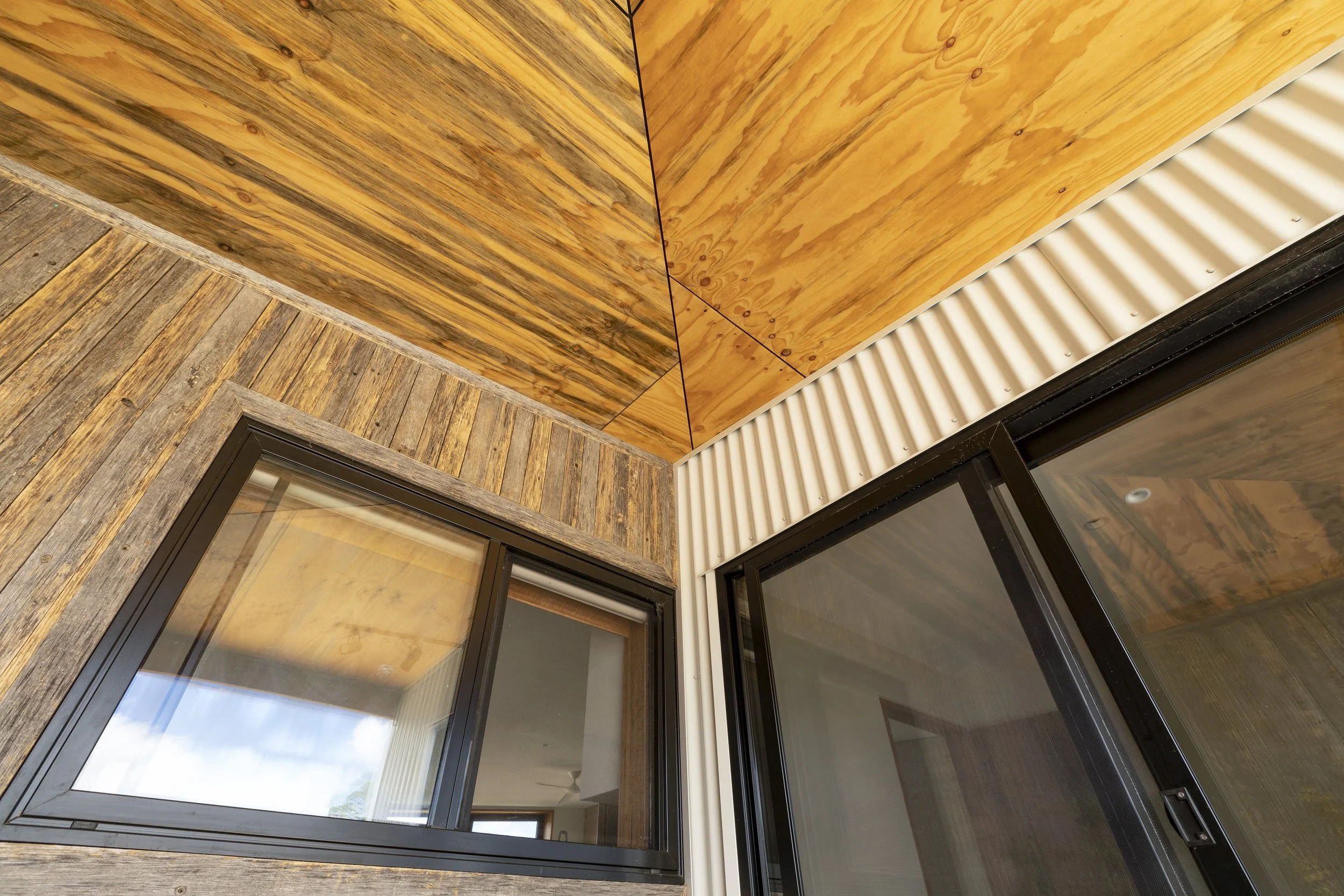 Close-up view of a corner of a building with a wooden exterior, showing large windows and a part of the roof with wooden paneling and metal siding.