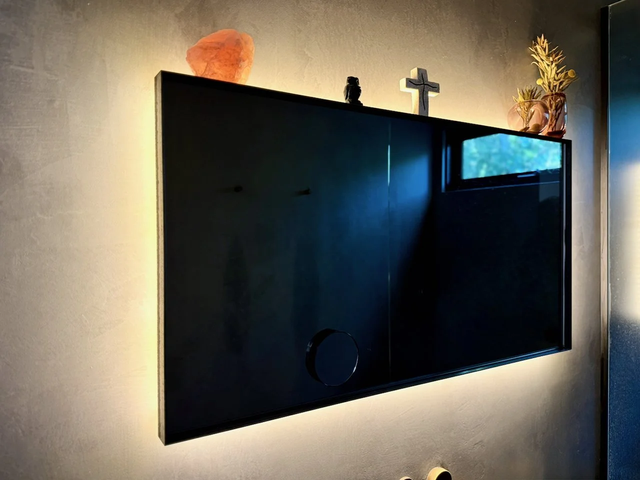 Black flat-screen TV mounted on a wall with backlit lighting, above which decorative items including a salt lamp, small statue, cross, and a glass vase with plants are placed.
