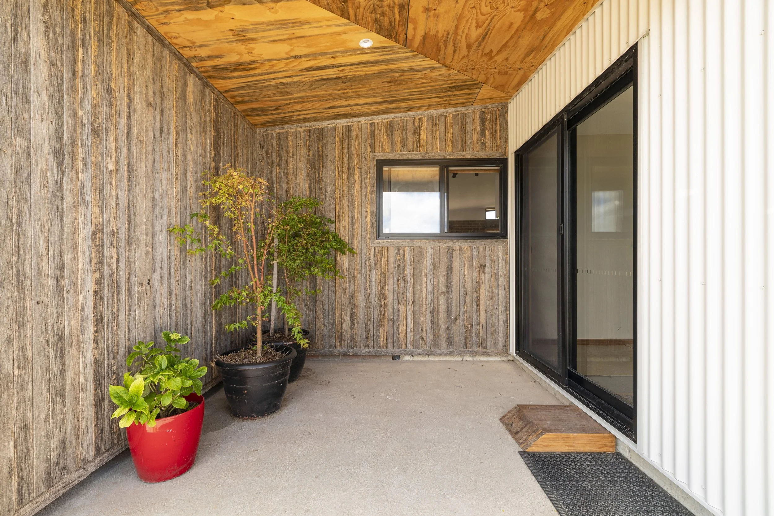 Small balcony with three potted plants, two large ones in black and red pots and a small green one in a red pot. The balcony has a wooden wall and ceiling, a sliding glass door with a black frame, and a beige corrugated metal wall.
