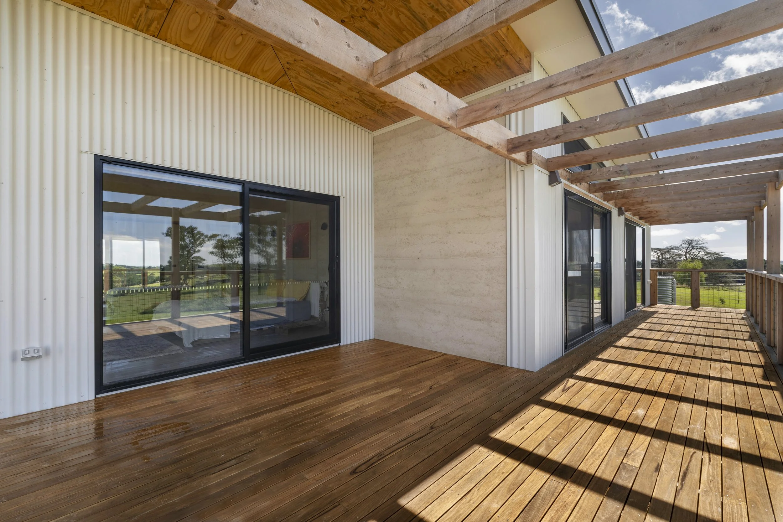 An outdoor balcony with wooden flooring, sliding glass doors, and a roof with exposed wooden beams. The landscape includes trees and a partly cloudy sky.