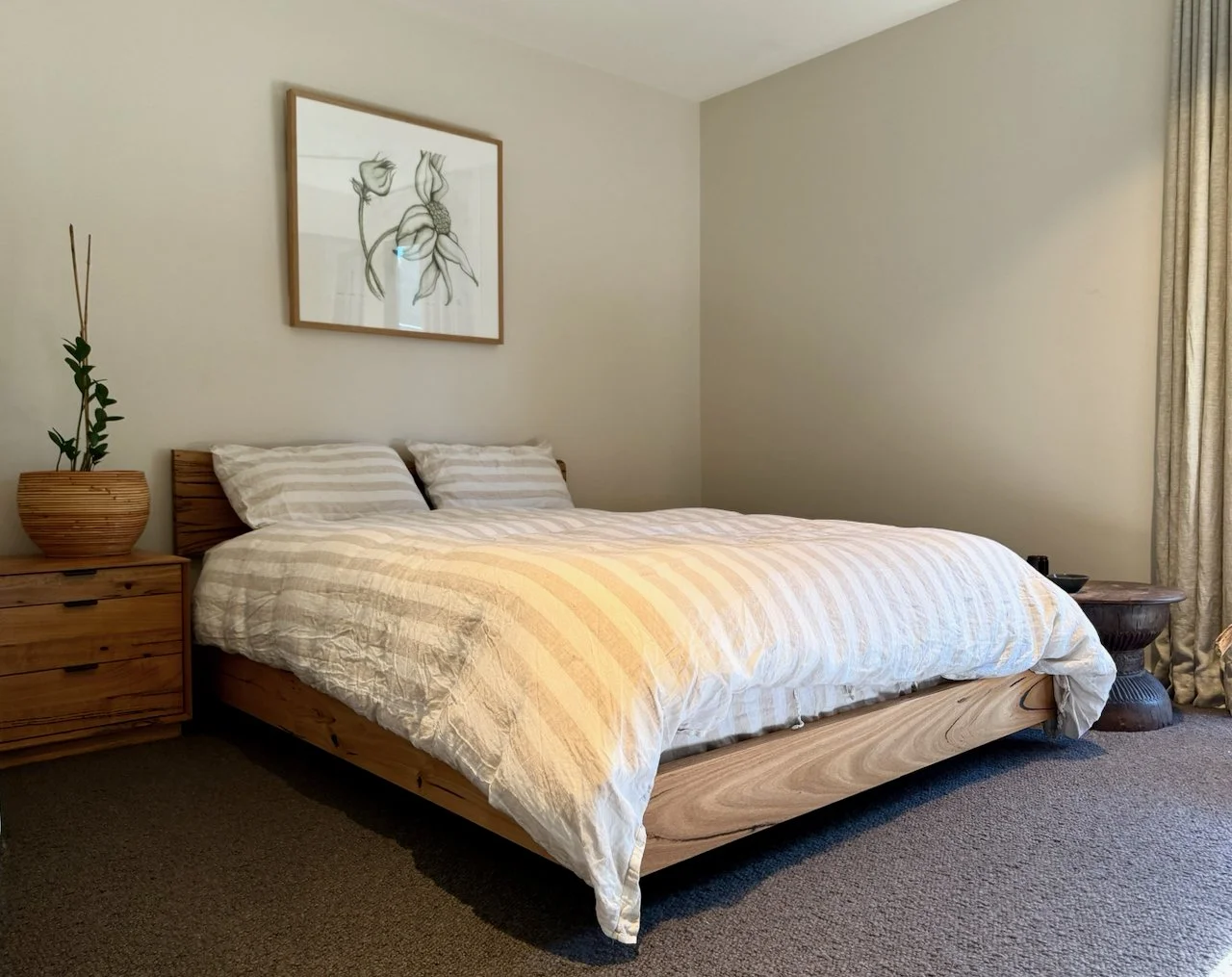 A minimalist bedroom with a wooden bed frame, a white striped duvet, and two pillows. There is a wooden nightstand with a plant in a woven pot on the left side and a round wooden side table with objects on the right. A framed black and white line dra