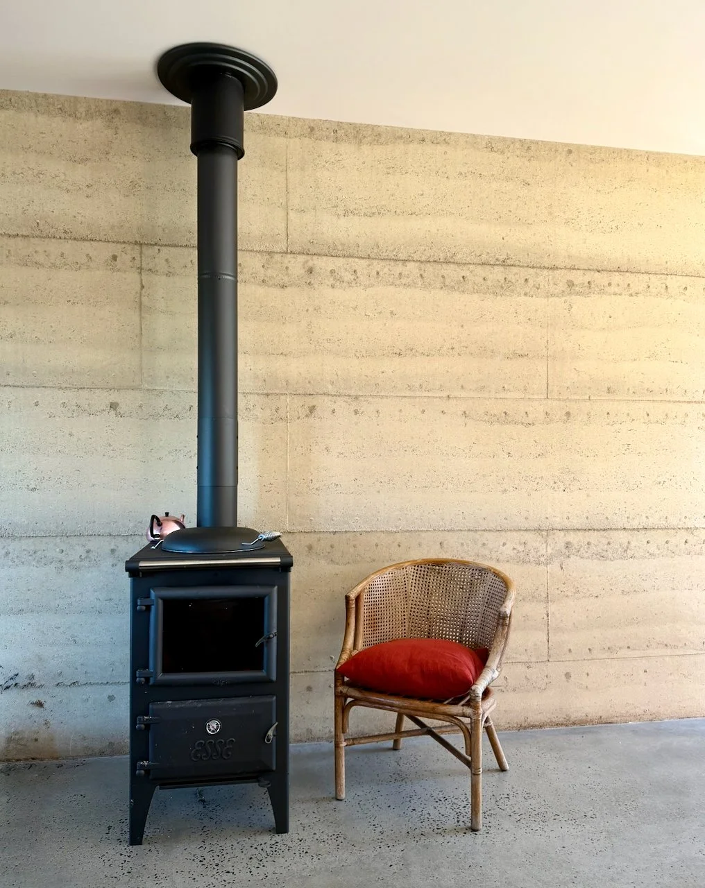 A black wood-burning stove with a pipe extending up to the ceiling, next to a wicker chair with a red cushion, against a concrete wall.
