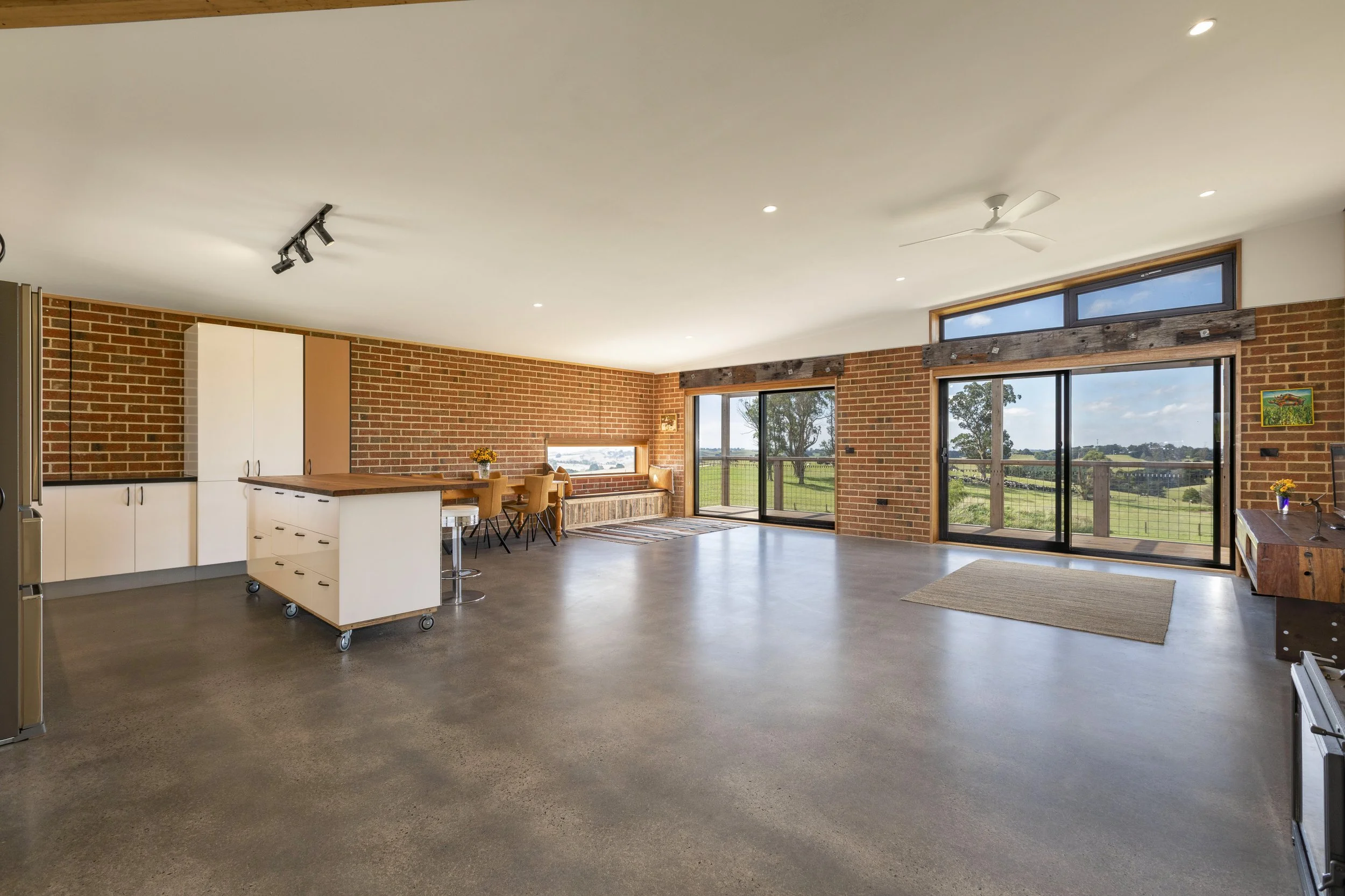 Interior view of a spacious living area with brick walls, large glass sliding doors, and a view of greenery outside, featuring a kitchen island, dining area, and ceiling fan.
